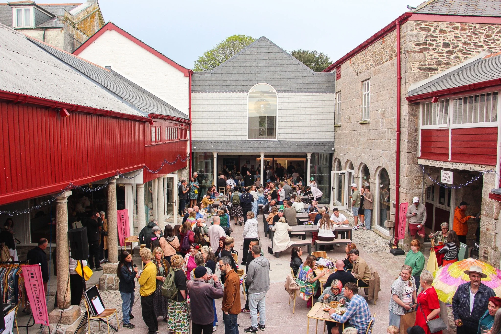 Crowd of people gathered in an outdoor courtyard surrounded by colorful historic buildings with red, white, and gray facades, some with stone walls, during daytime.