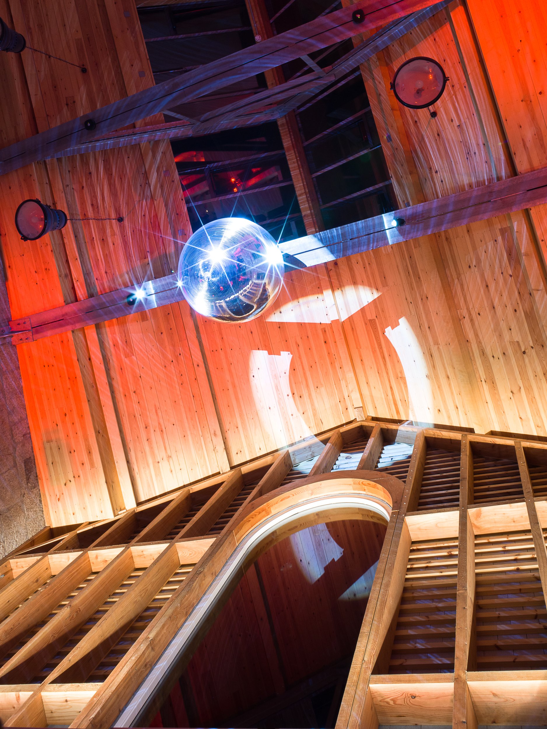 Wooden spiral staircase with glass railing, colorful lights, and a disco ball reflecting light on the ceiling.