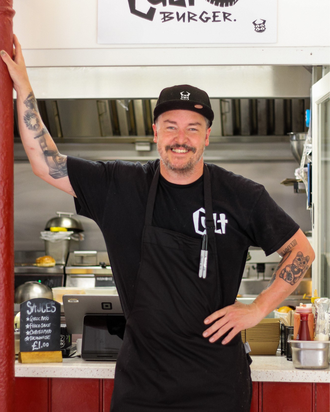 Smiling man with tattoos wearing a black cap and black apron standing behind a counter in a fast-food kitchen, with condiments and a cash register visible in the background.