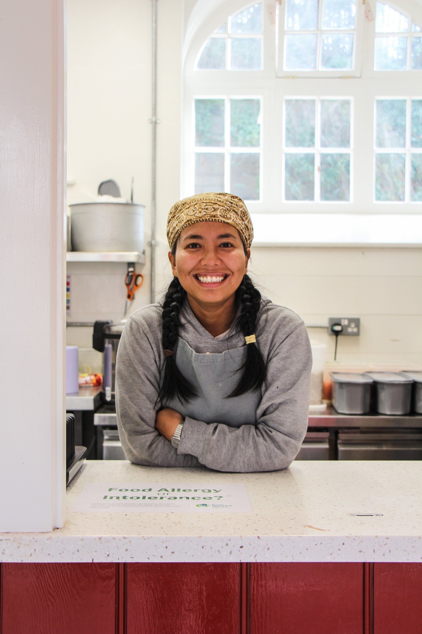Smiling woman with dark hair in braids, wearing a bandana and apron, standing behind a food allergy information sign on a counter in a kitchen or food service setting.