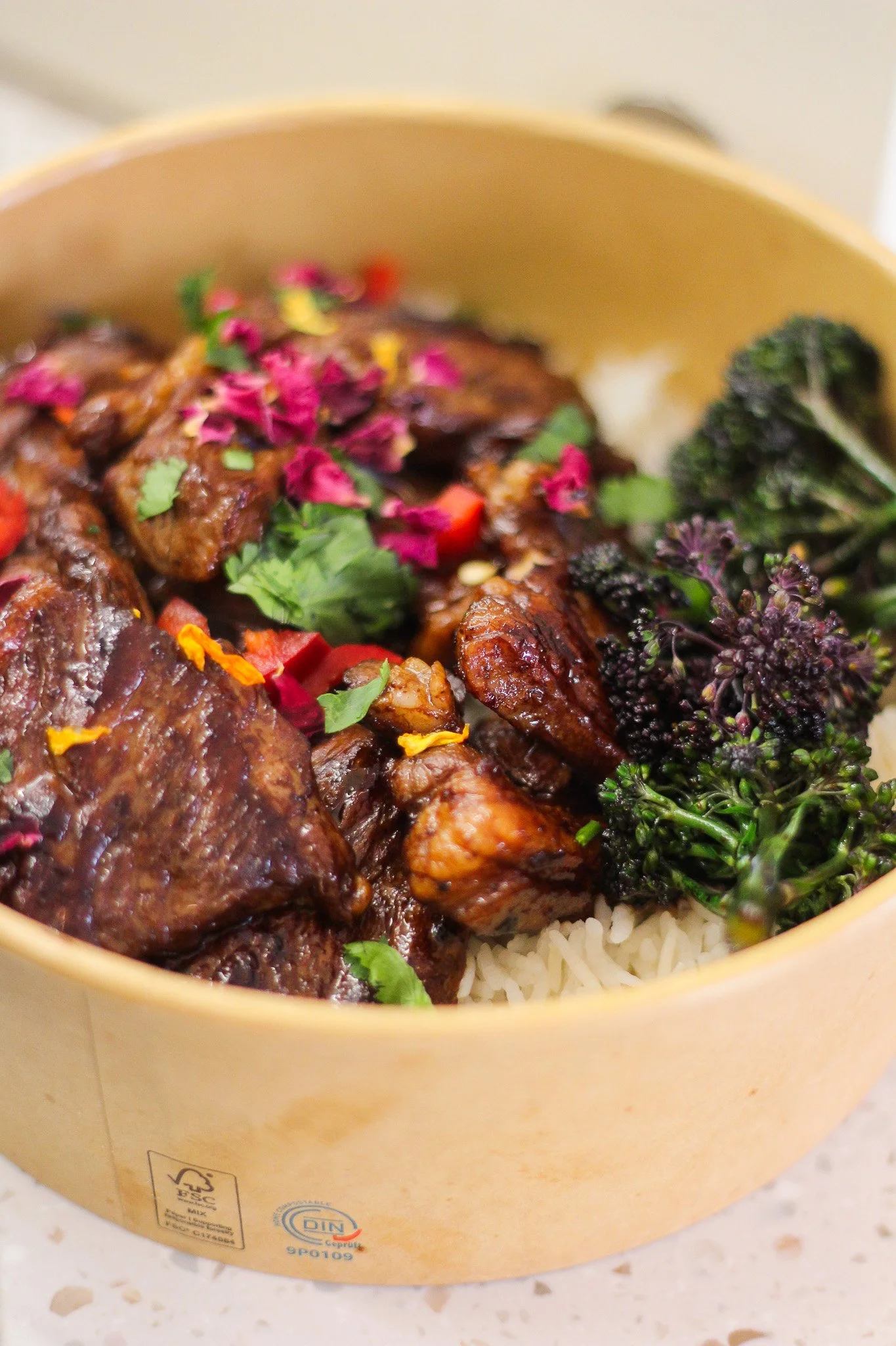 A bowl of rice with grilled meat, garnished with herbs and flowers, and served on a white surface.
