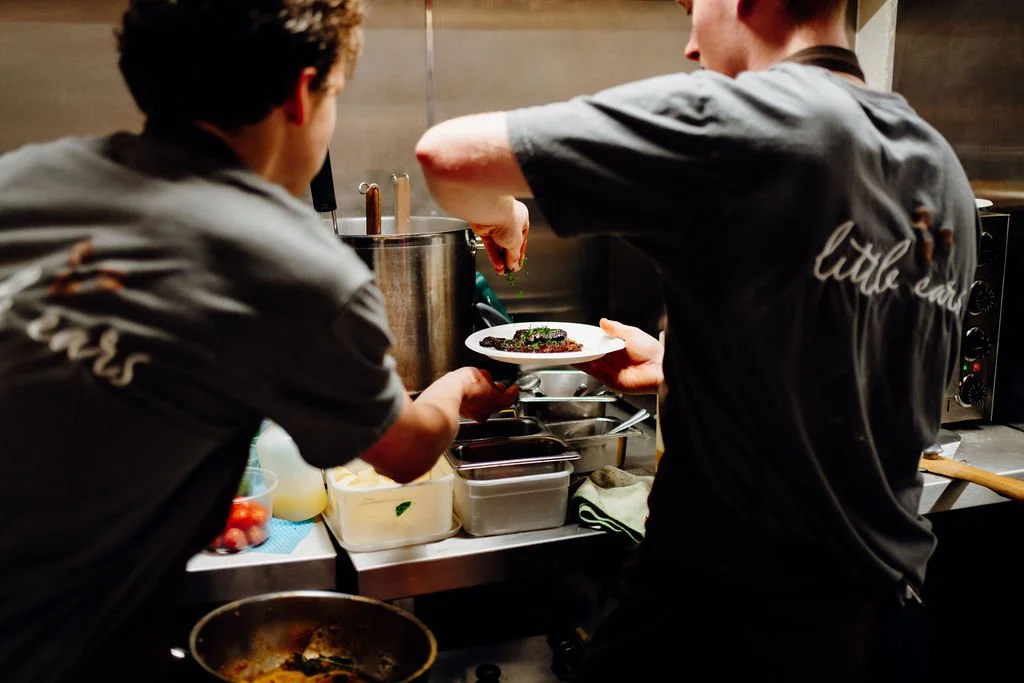 Two chefs back to back preparing food in a commercial kitchen with various ingredients and utensils on the counter.