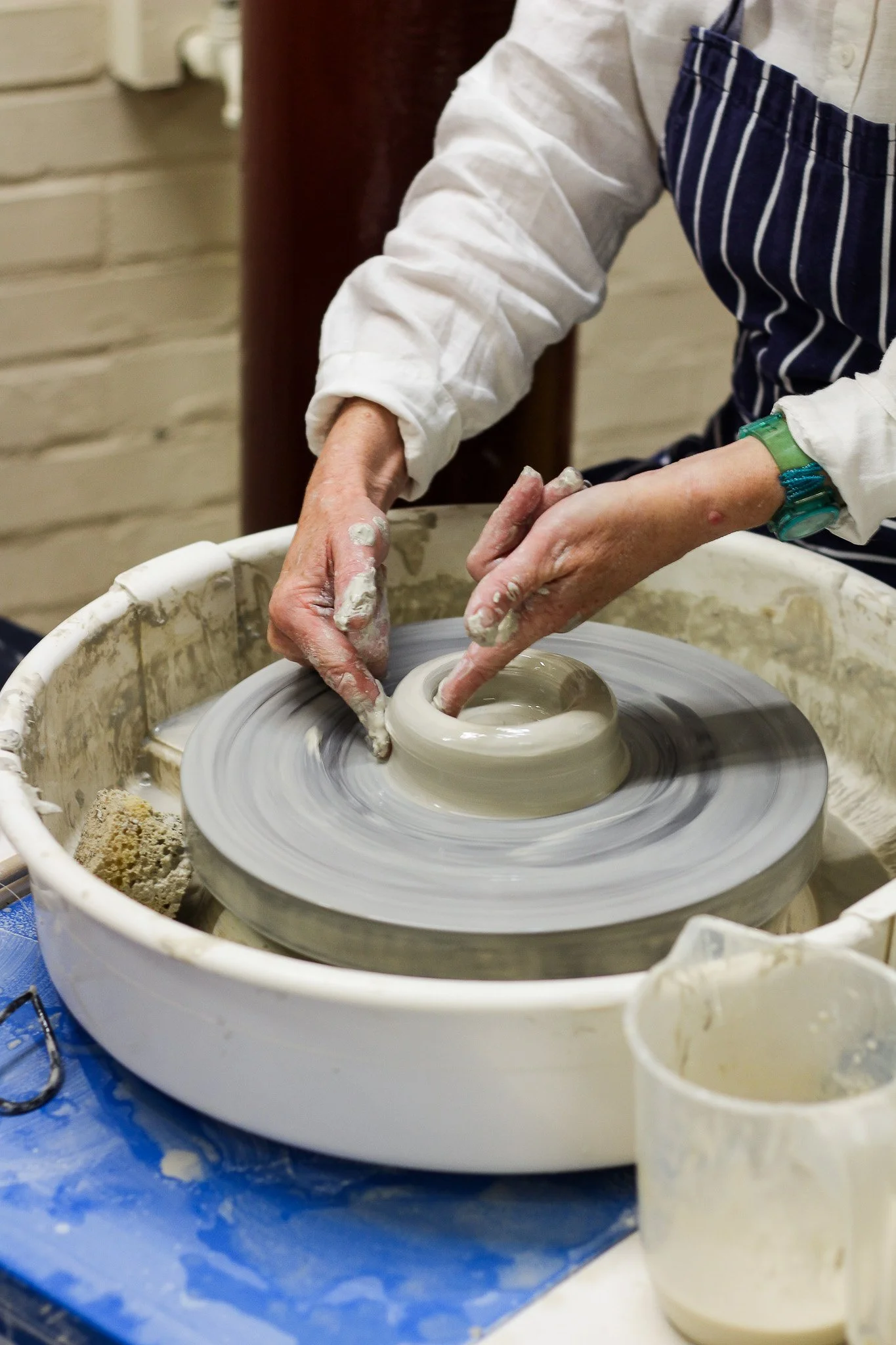 Person shaping clay on a pottery wheel in a studio, wearing a striped apron and a white long-sleeve shirt.