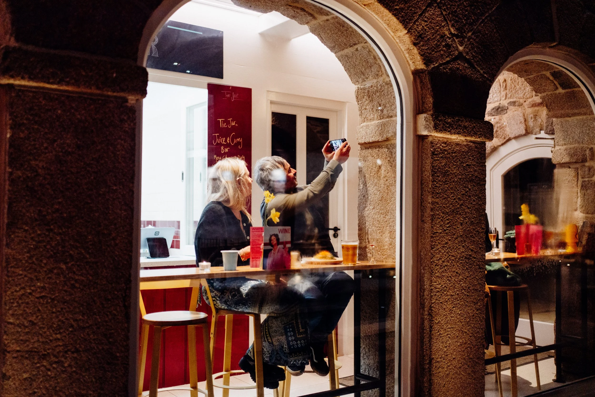 View through a window showing two women inside a restaurant or cafe. One woman is taking a selfie with her phone, while the other looks on. The interior has a red sign, a TV, and a window at the back.