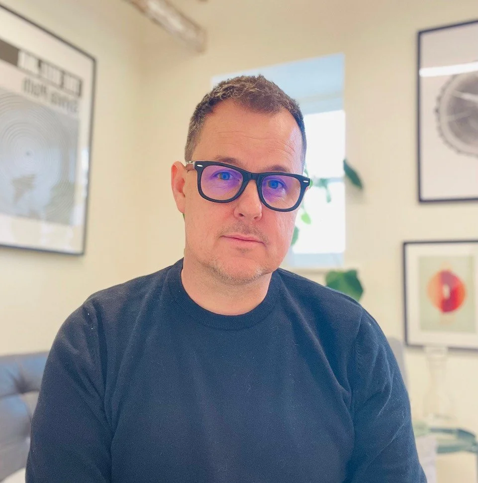 Man with short dark hair, glasses, wearing a black shirt, sitting in a room with framed artwork on the walls, a window with sunlight, and a plant in the background.