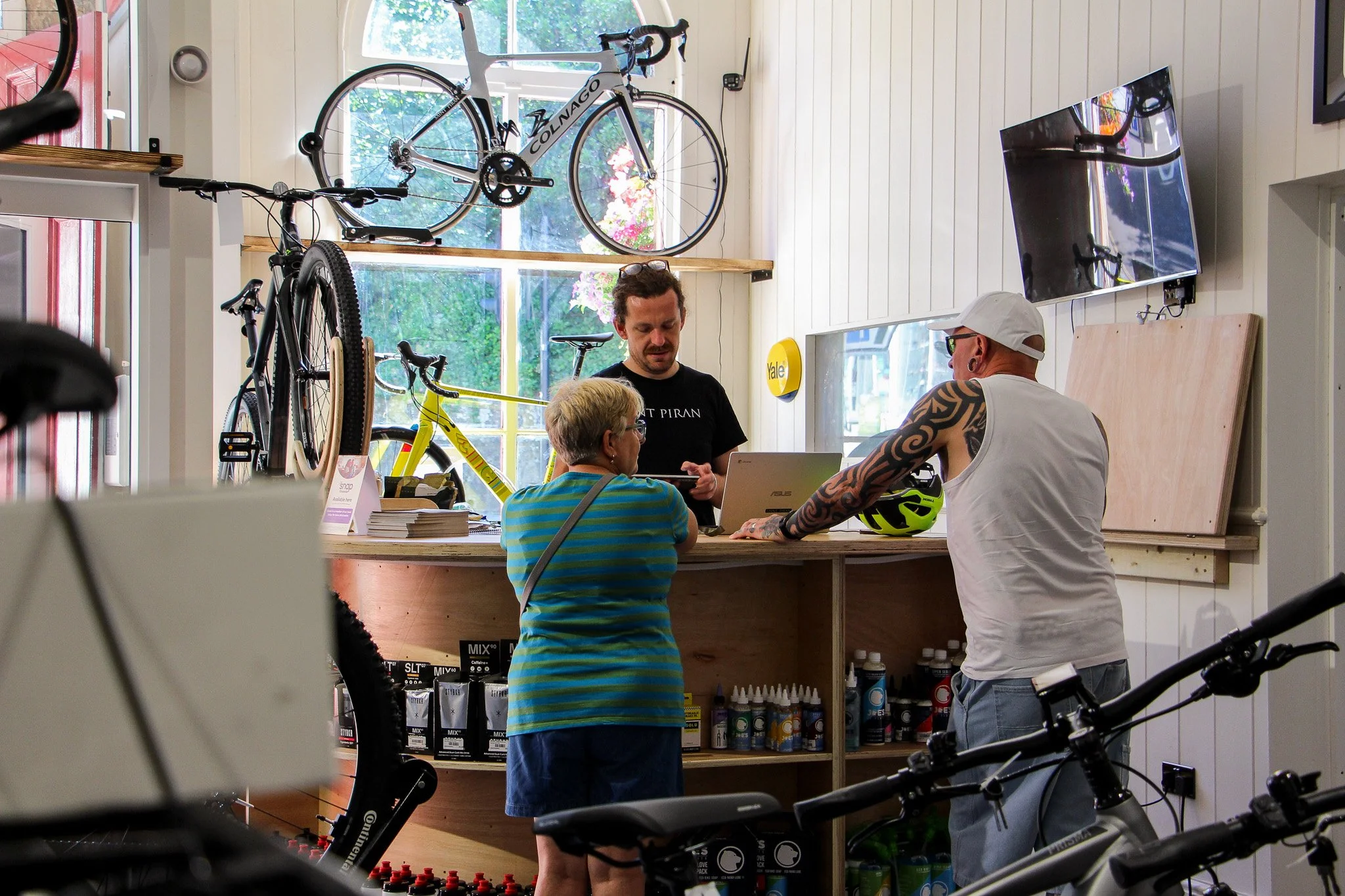 Three people standing at a counter inside a shop, engaging in conversation. Bicycles and cycling gear are visible, with a bicycle mounted on the wall and others on the floor. Shelves behind them display various bottles and products. Large windows with trees outside are in the background.