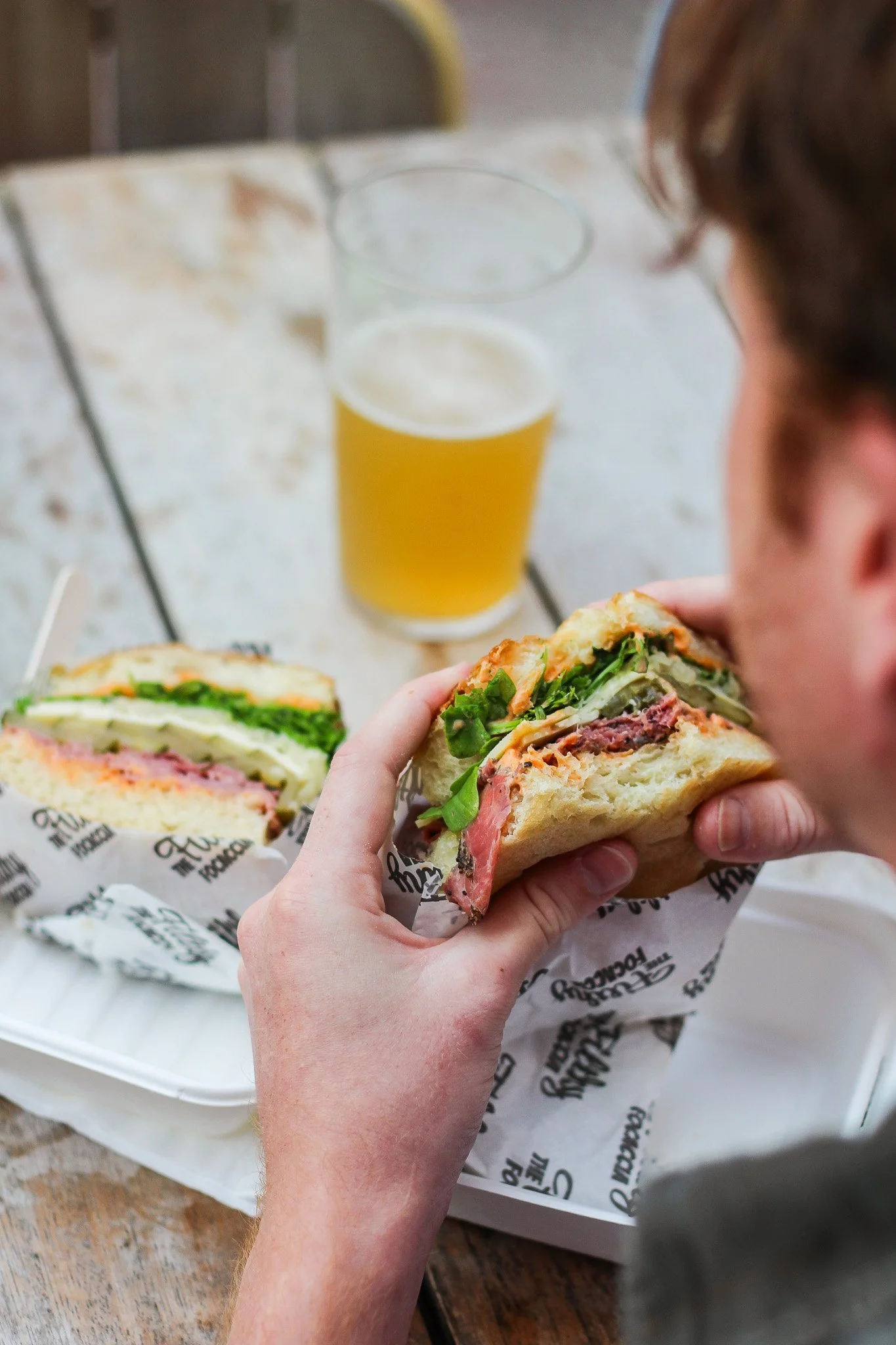 Close-up of a person holding a sandwich with lettuce, meat, and other fillings. In the background, there is a cup of beer on a table and a sandwich wrapped in paper. The scene takes place in a casual dining setting.