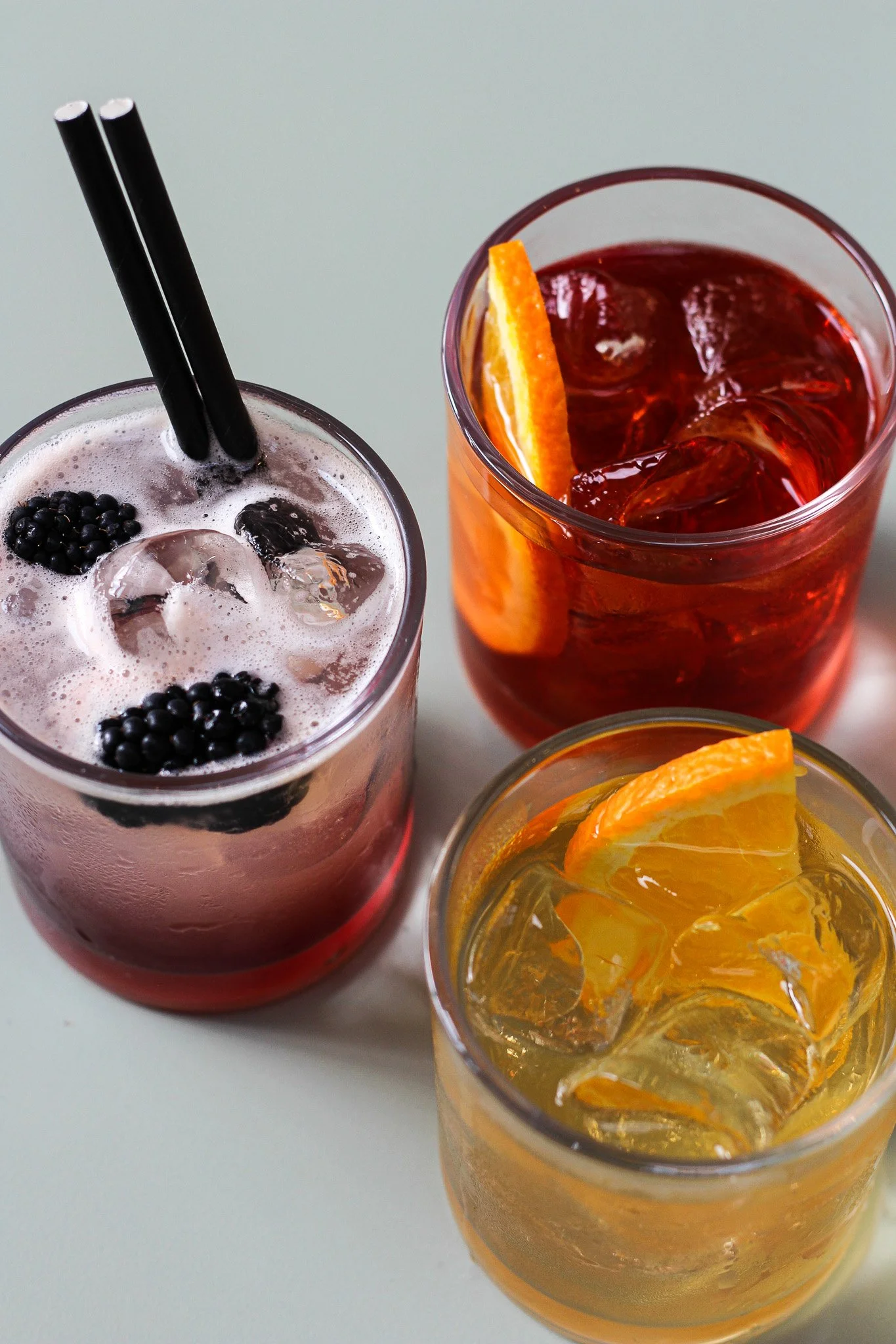 Three glasses of colorful drinks with slices of orange and ice, viewed from above on a white surface.