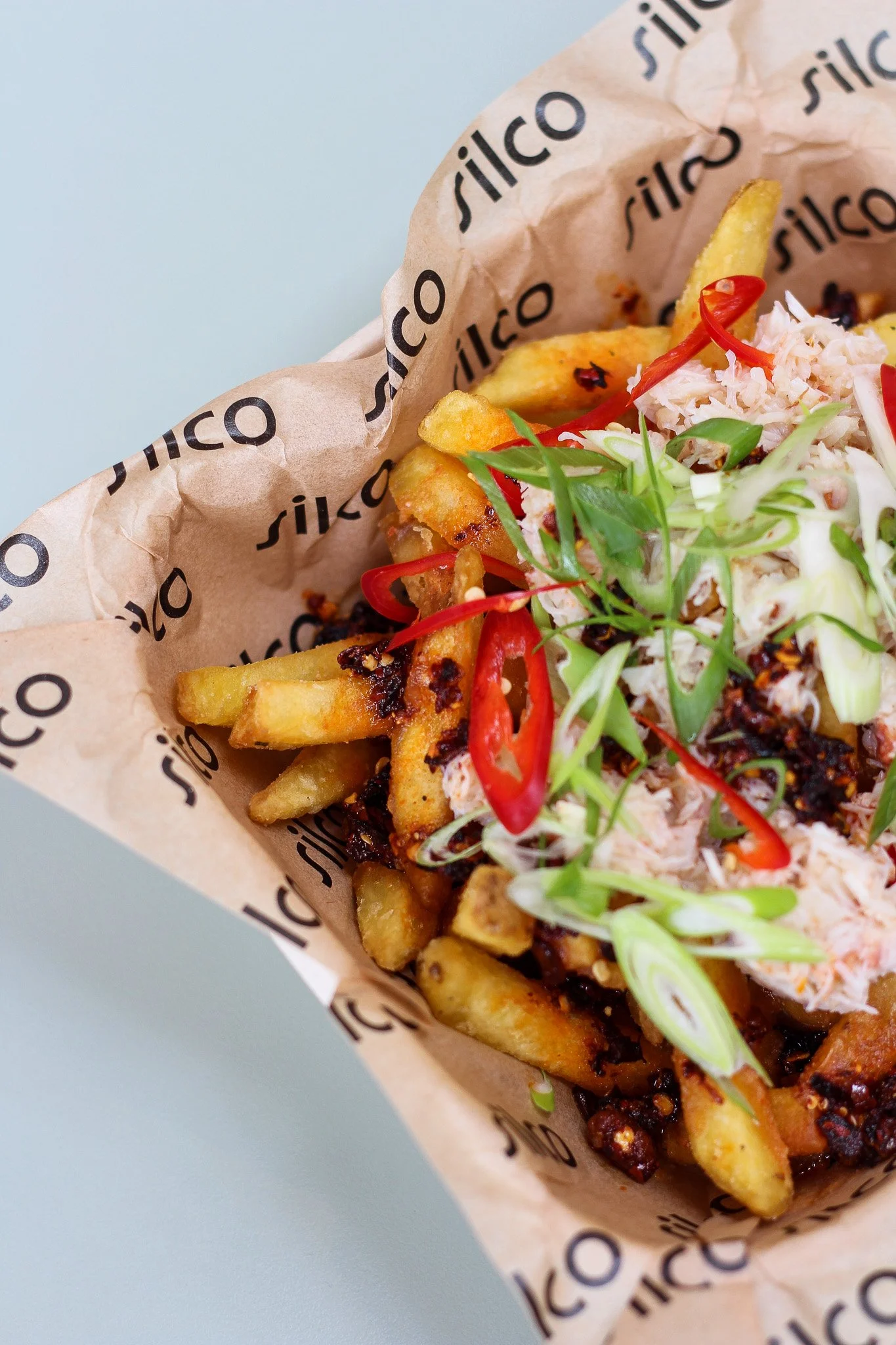 Close-up of loaded fries and a side of shredded chicken topped with sliced red chili, green onion, and sauce in a paper serving tray with branding.