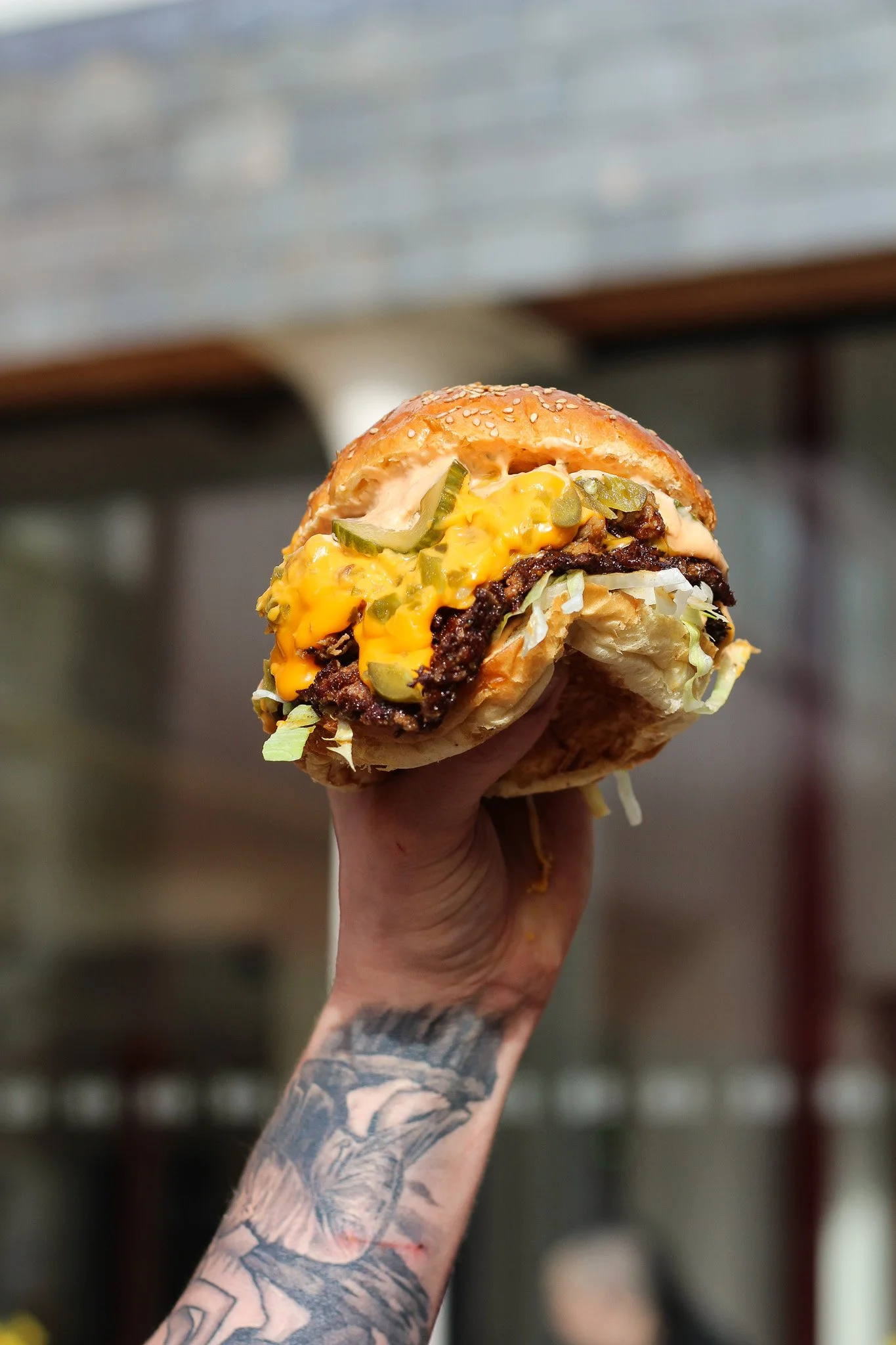 Close-up of a hand holding a cheeseburger with pickles, lettuce, cheese, and a beef patty in a sesame seed bun.