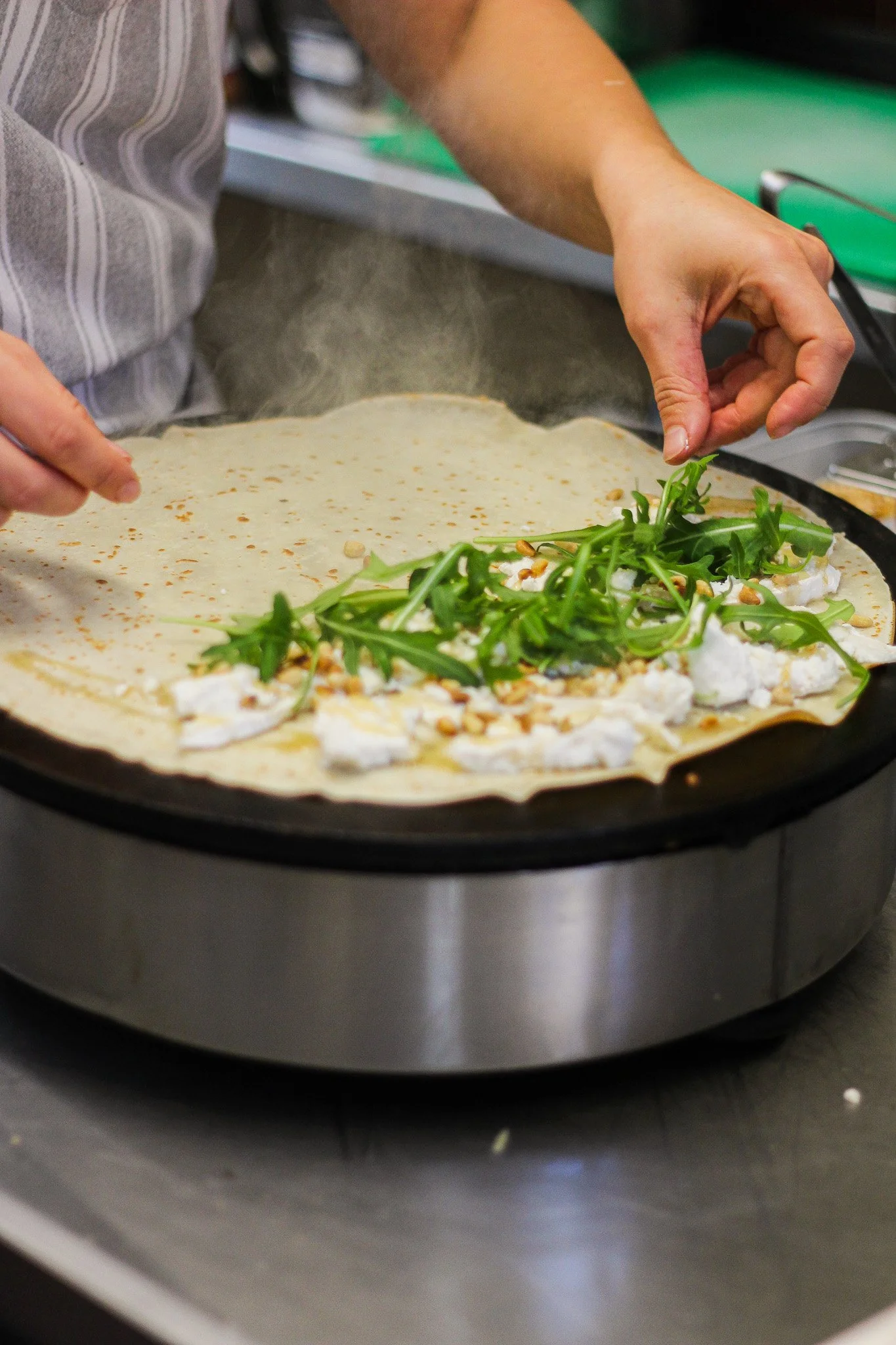 Hands preparing a pizza with toppings of arugula, cheese, and pine nuts inside a pizza oven.