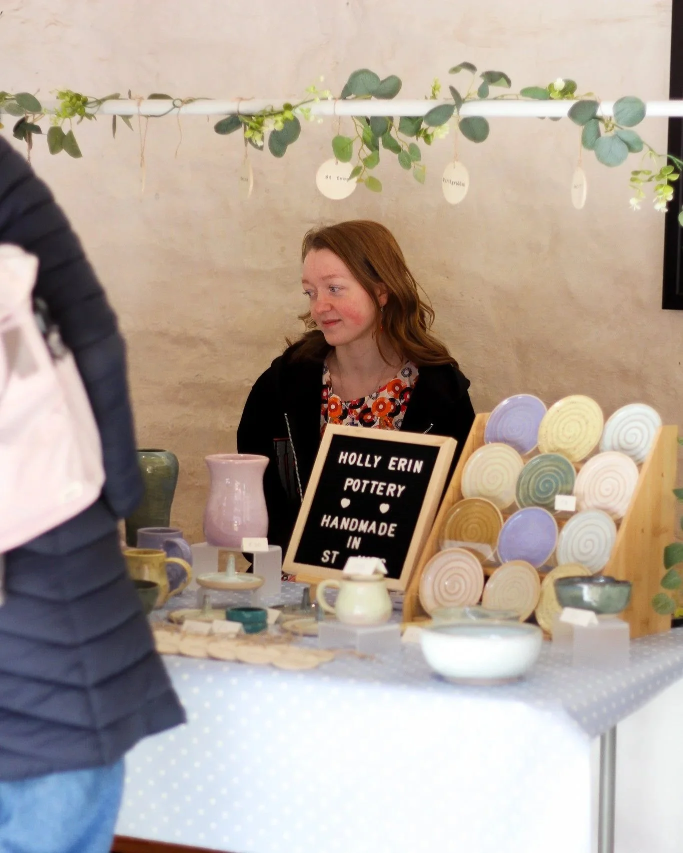 A woman with red hair sitting at a table selling handmade pottery at a craft fair. The table displays various ceramic bowls, mugs, and cups with a sign that reads 'Holly Erin Pottery Handmade in St'. A person in a black jacket is standing in front of the table. There is a decorative green vine hanging above the booth.