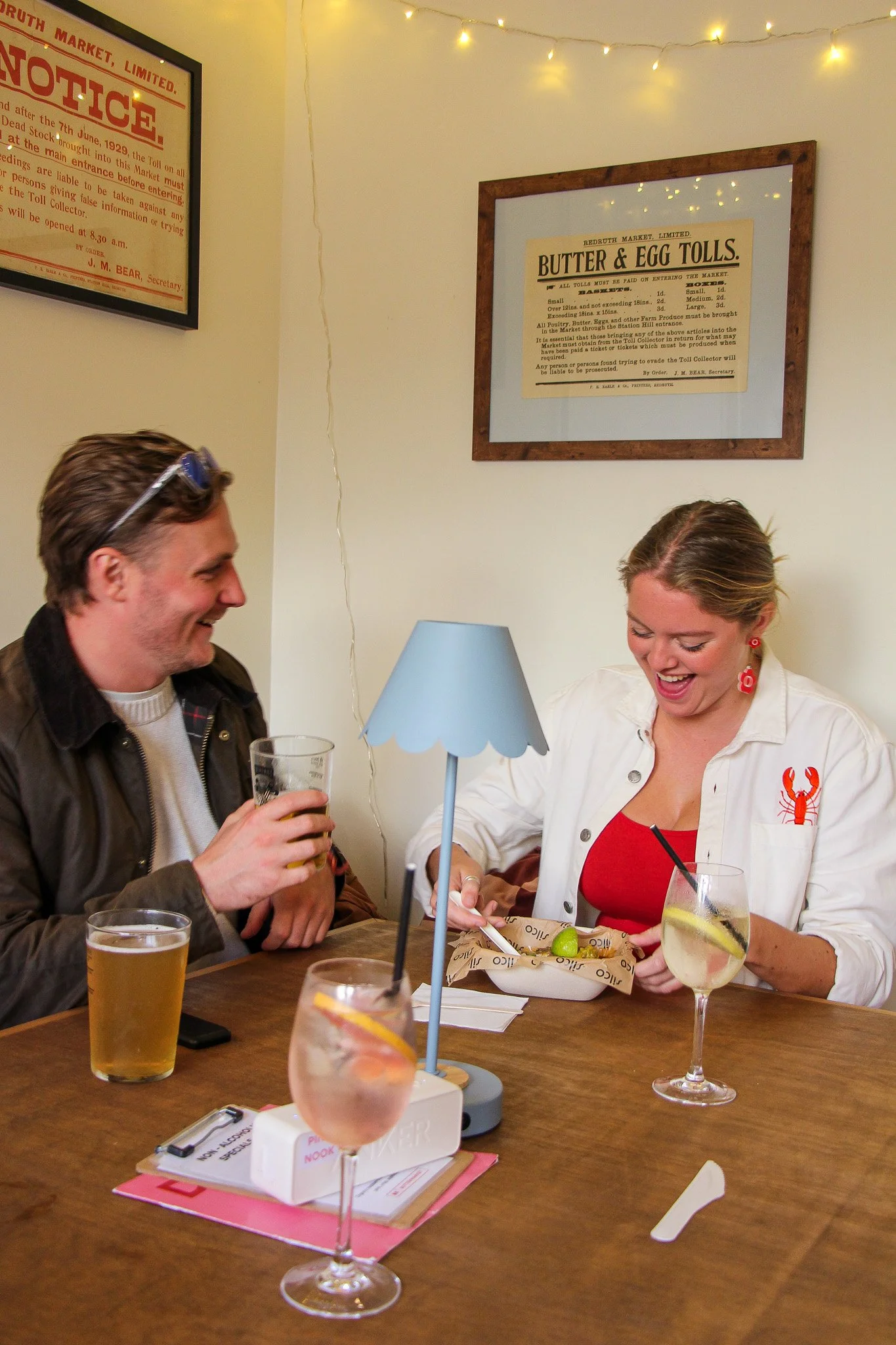 Two people sitting at a wooden table enjoying drinks and food at a restaurant or pub. The man on the left is holding a pint of beer and smiling, while the woman on the right is looking down at a plate of food, smiling. There are various drinks and a tablet on the table, and a framed notice on the wall behind them. The setting appears cozy with soft lighting and decorative string lights.