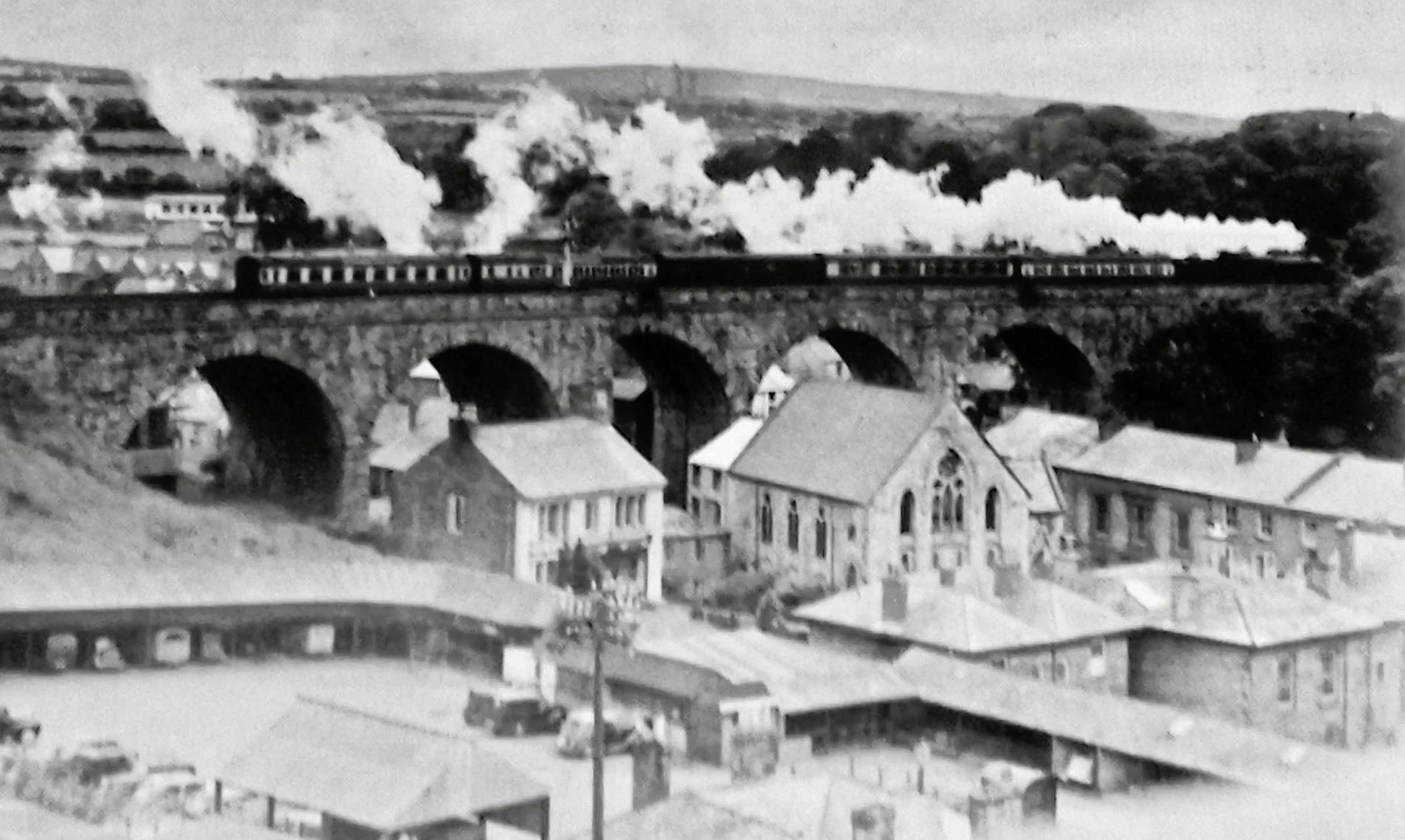 Black and white photograph of a steam train crossing a stone arch bridge over a small town with houses and buildings in the foreground, smoke billowing from the train's engines, and rolling hills in the background.