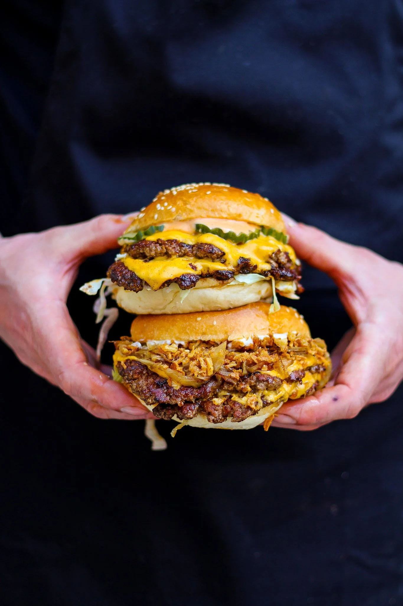 Close-up of a person holding a large cheeseburger with multiple layers of beef patties, cheese, pickles, and a bun against a dark background.