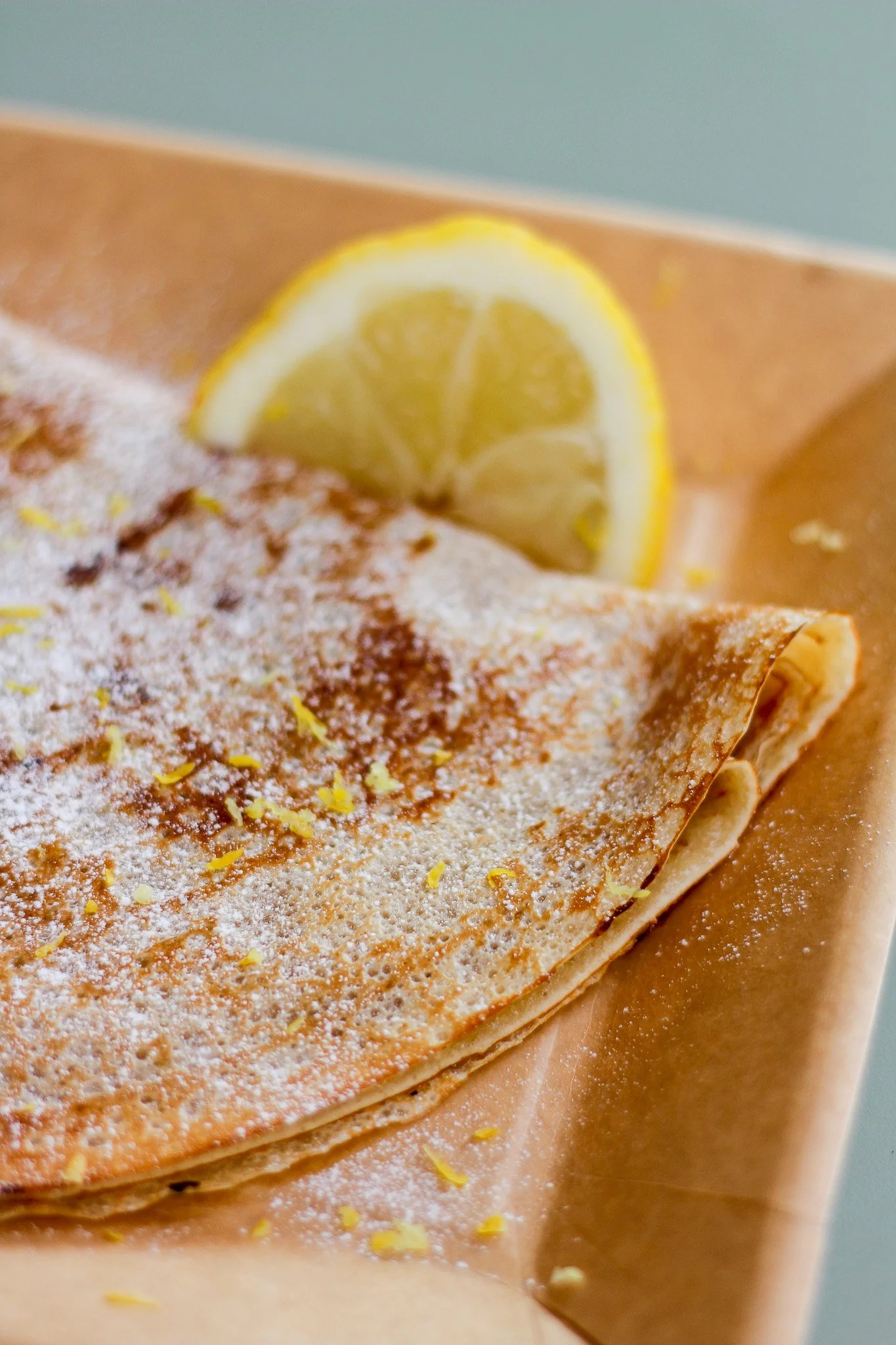 Close-up of a lemon slice on a dessert with powdered sugar and lemon zest, placed on parchment paper.
