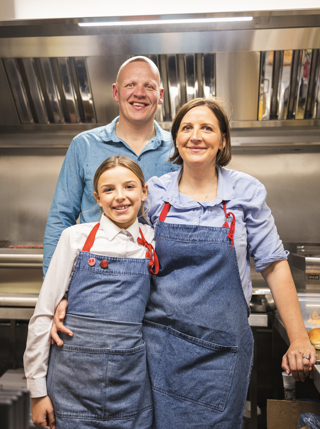 A family of three, including a man, woman, and girl, standing together in a commercial kitchen, smiling at the camera. They are wearing matching blue aprons, indicating they are working together, possibly in a culinary or restaurant setting.