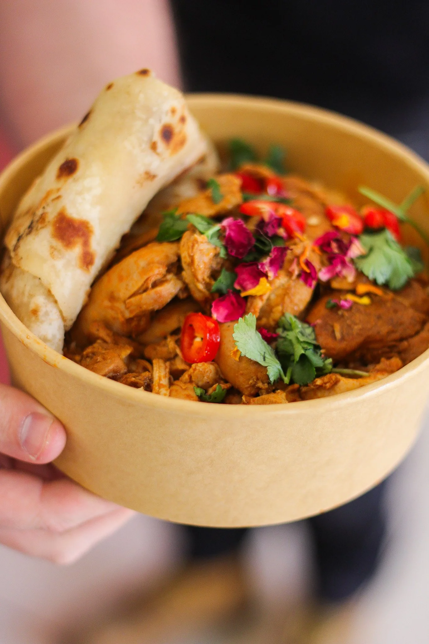 A person holding a beige bowl filled with Chinese takeout food, including a spring roll, fried chicken, and garnished with cilantro, red chili slices, and edible flower petals.
