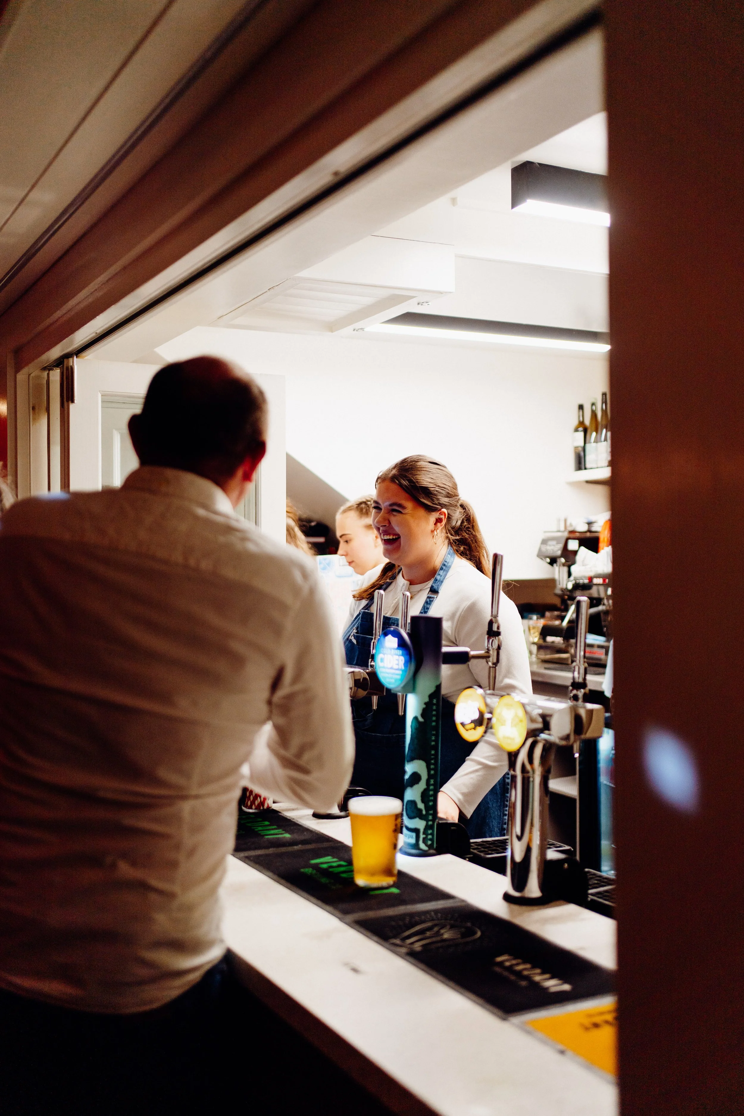 A woman with brown hair, wearing a white shirt and blue apron, serving beer to a customer at a bar or pub. The customer has short hair and is wearing a beige shirt. There are beer taps, glasses, and bottles in the background, with bright lighting and a white interior.