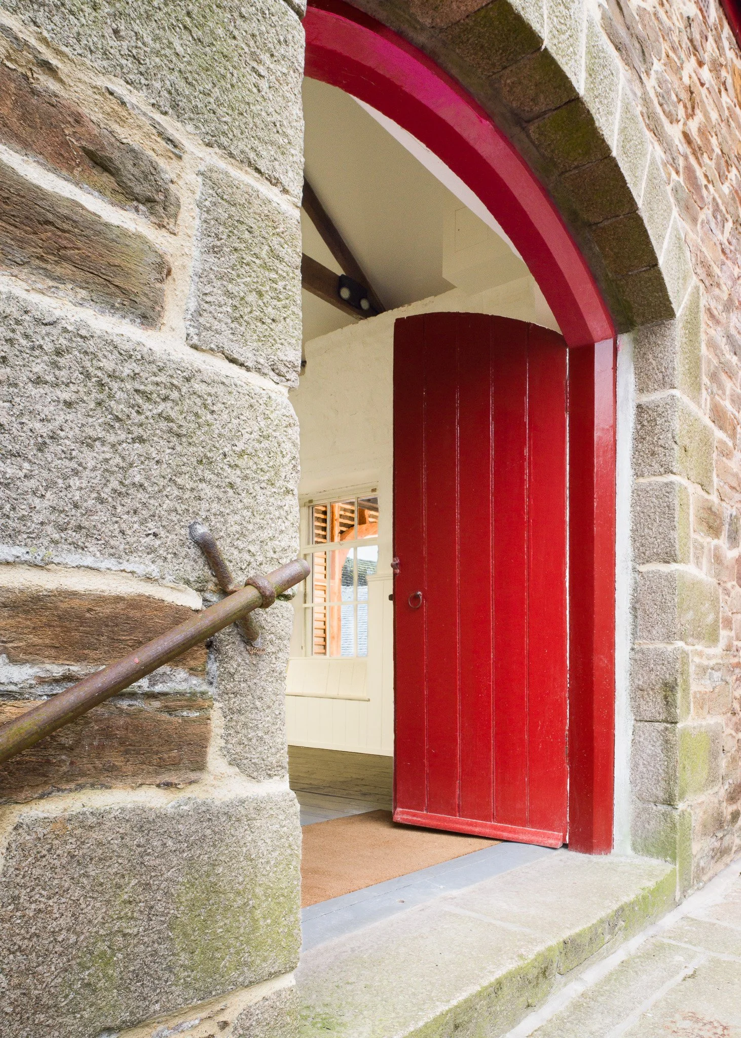 A red wooden door partially open, framed by stone walls and an arch, revealing a glimpse of the interior with a white window and wooden staircase.