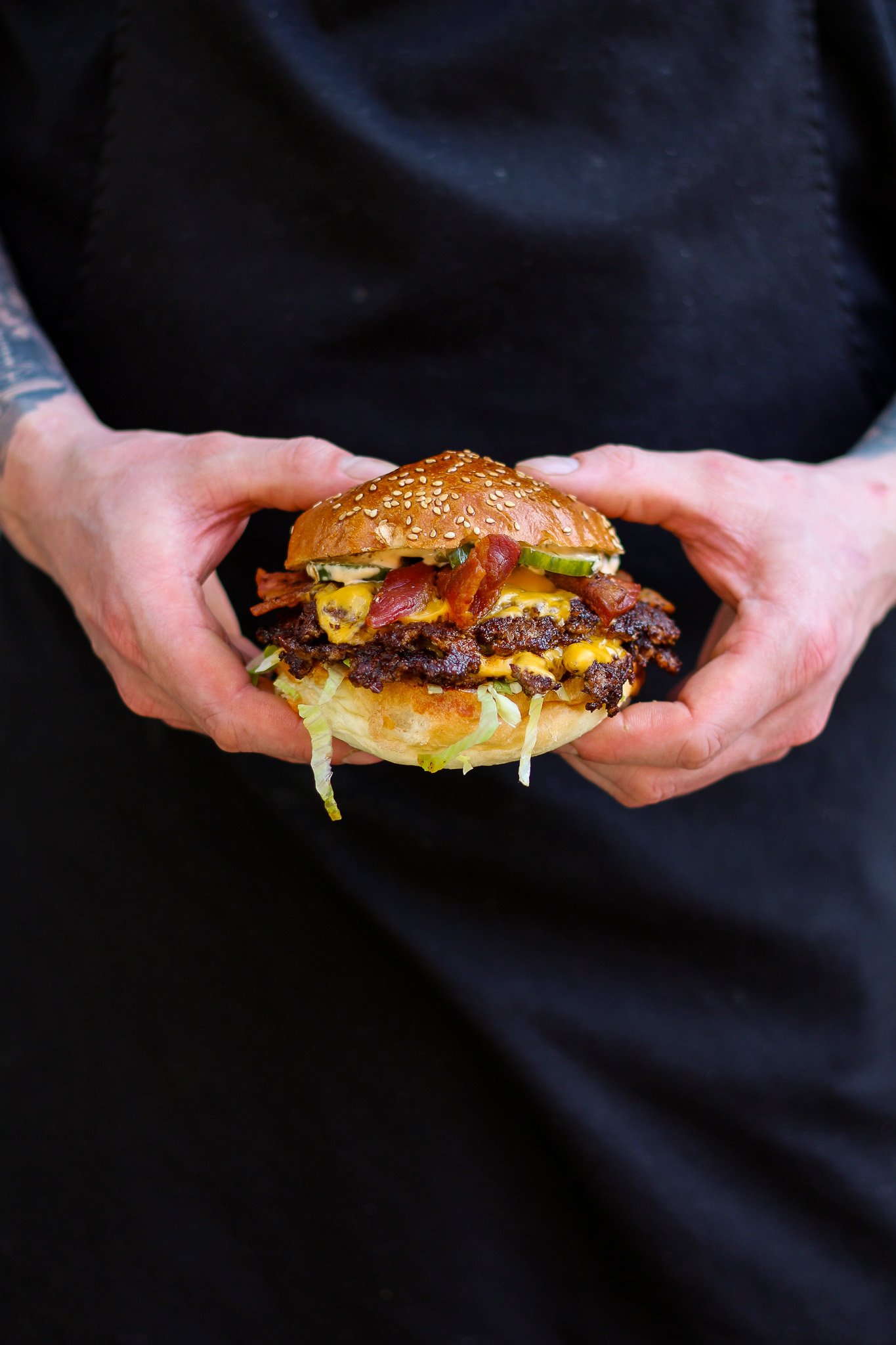 Person holding a large cheeseburger with lettuce, pickles, bacon, and sesame seed bun against a black background.
