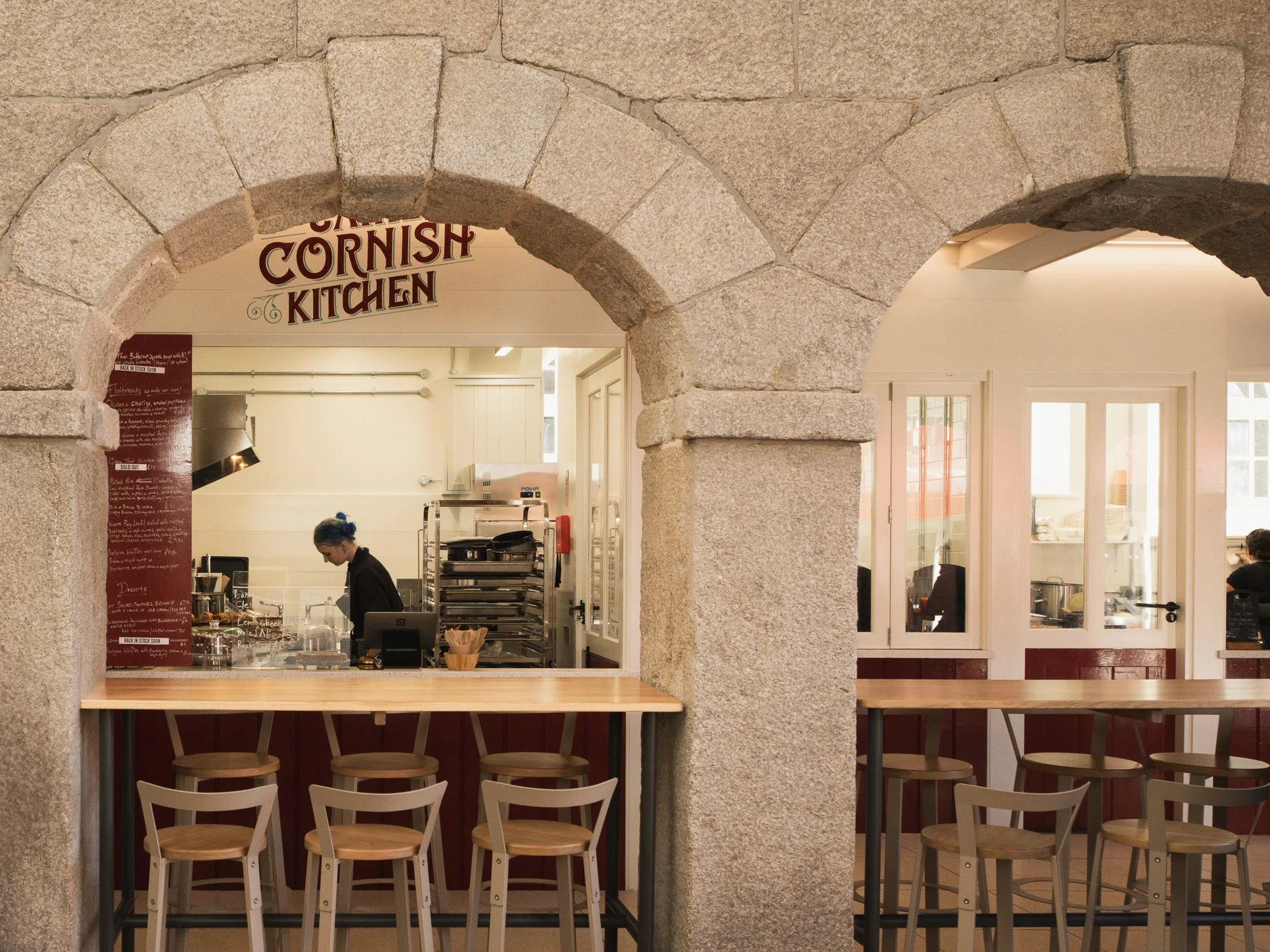 Inside a restaurant called Cornish Kitchen, visible through an open stone archway, with a counter and staff preparing food, and seating area with chairs.
