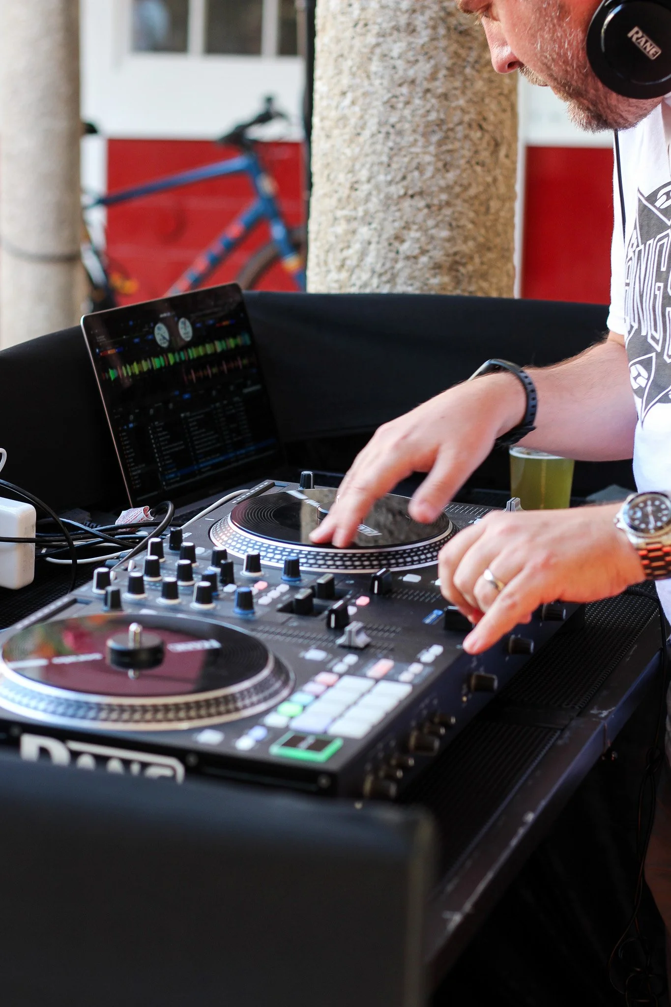 A man wearing headphones and a watch, focused on DJ equipment with turntables and a laptop, outdoors near a building with a bicycle in the background.