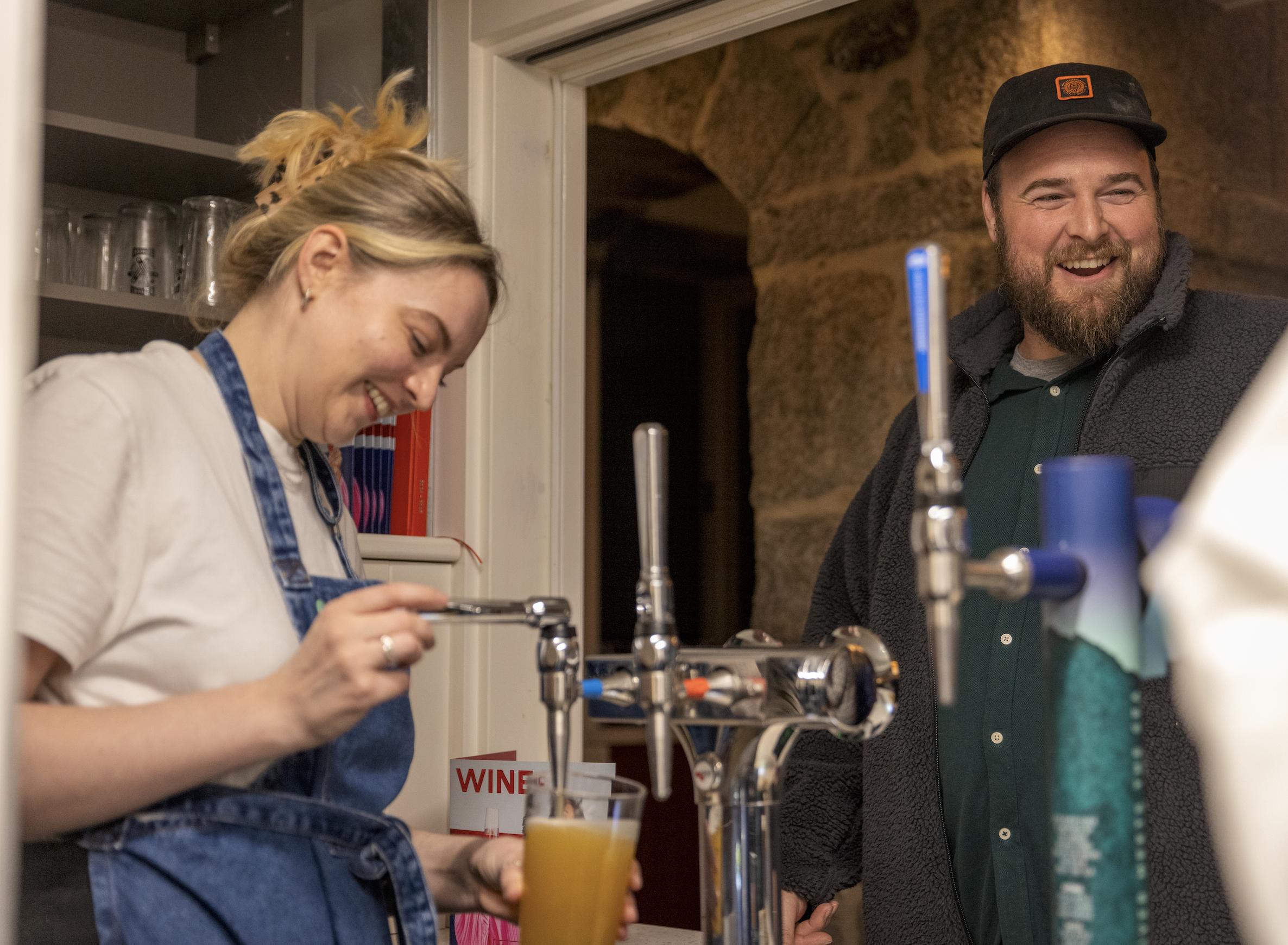 Two adults, a woman and a man, standing next to each other and smiling in a kitchen. The woman is pouring a drink into a glass, and the man, wearing a baseball cap, is smiling broadly. There are kitchen utensils and a sign that reads 'WINE' visible on the counter.