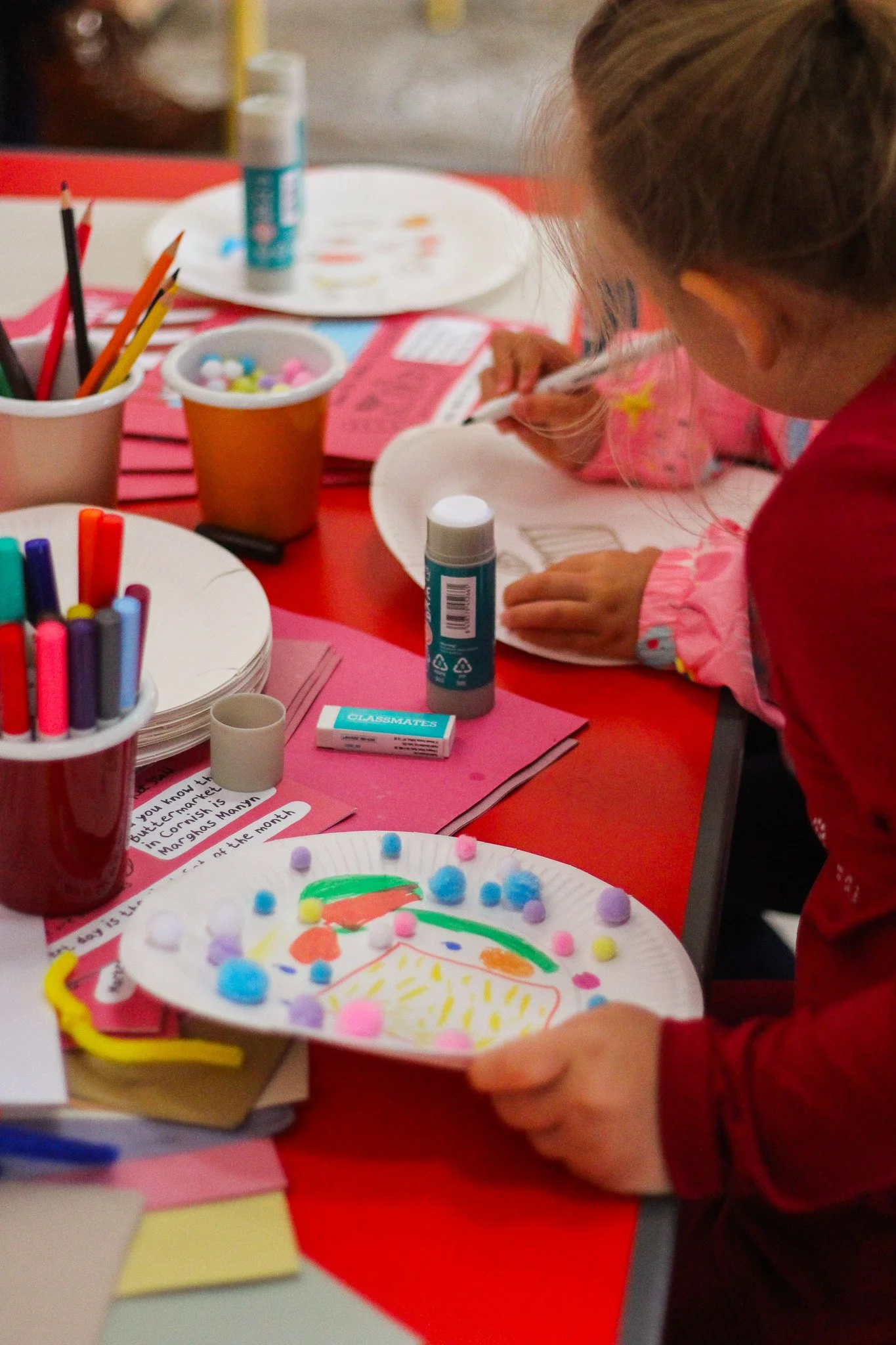 A young girl in a pink sweater and hair tied back at a craft table, decorating a paper plate with colorful pompoms and markers, surrounded by craft supplies including glue sticks, markers, and papers.