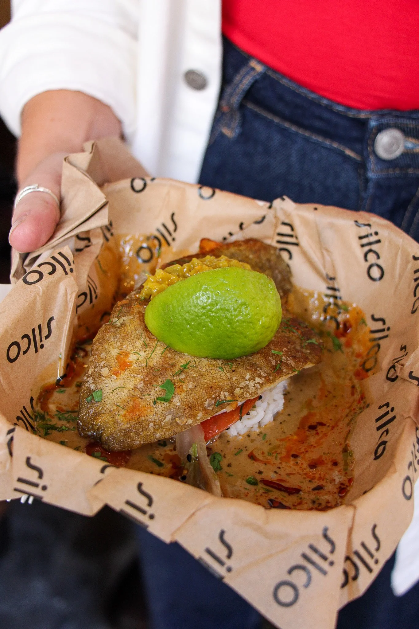 A fish taco with fried fish, rice, and toppings topped with a lime wedge, wrapped in branded paper.