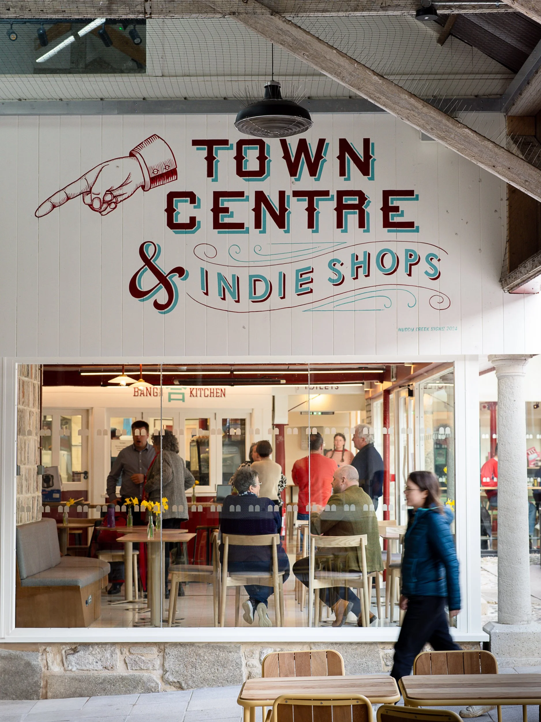 The image shows the interior of a busy cafe or restaurant viewed through a large glass window with people sitting at tables inside. The sign on the window reads 'TOWN CENTRE & Indie Shops,' and there is a hand pointing to the left next to the text. Outside, a person is walking past the window, and there are empty tables and chairs visible in the foreground.