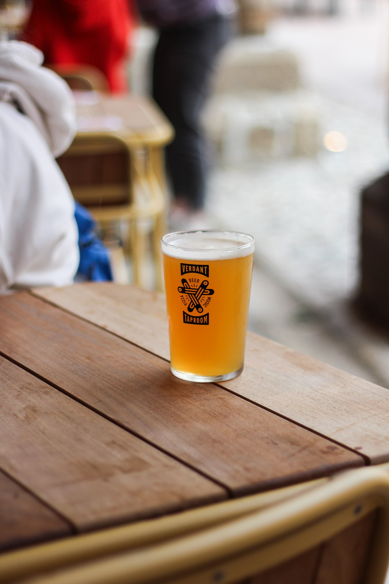 A glass of beer on a wooden table inside a bar or brewery, with a label that says "Verdant Beer Taproom." Blurred background of people and seating.