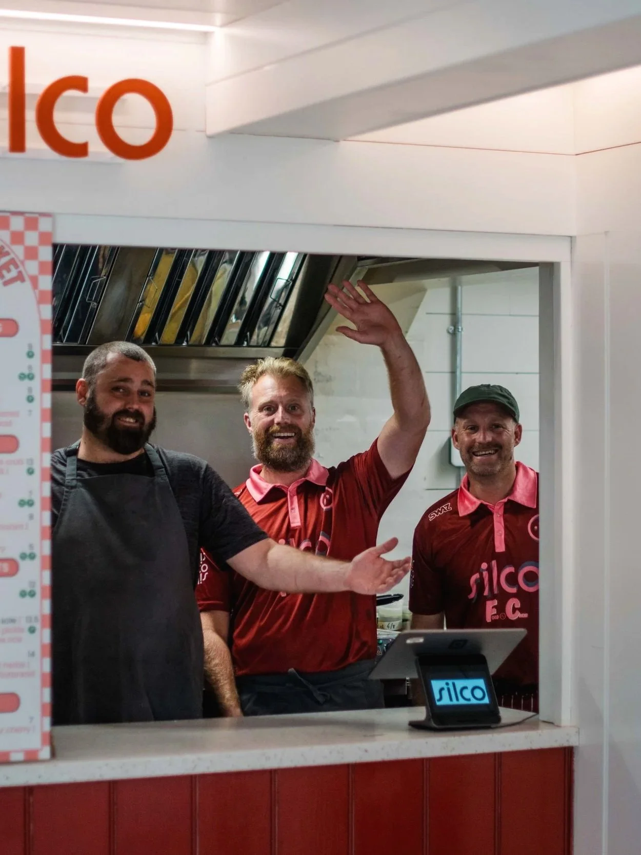 Three men smiling in a foodservice window, one wearing a black apron and two in red sports jerseys, with a sign reading 'silco' on a small digital device in front of them.