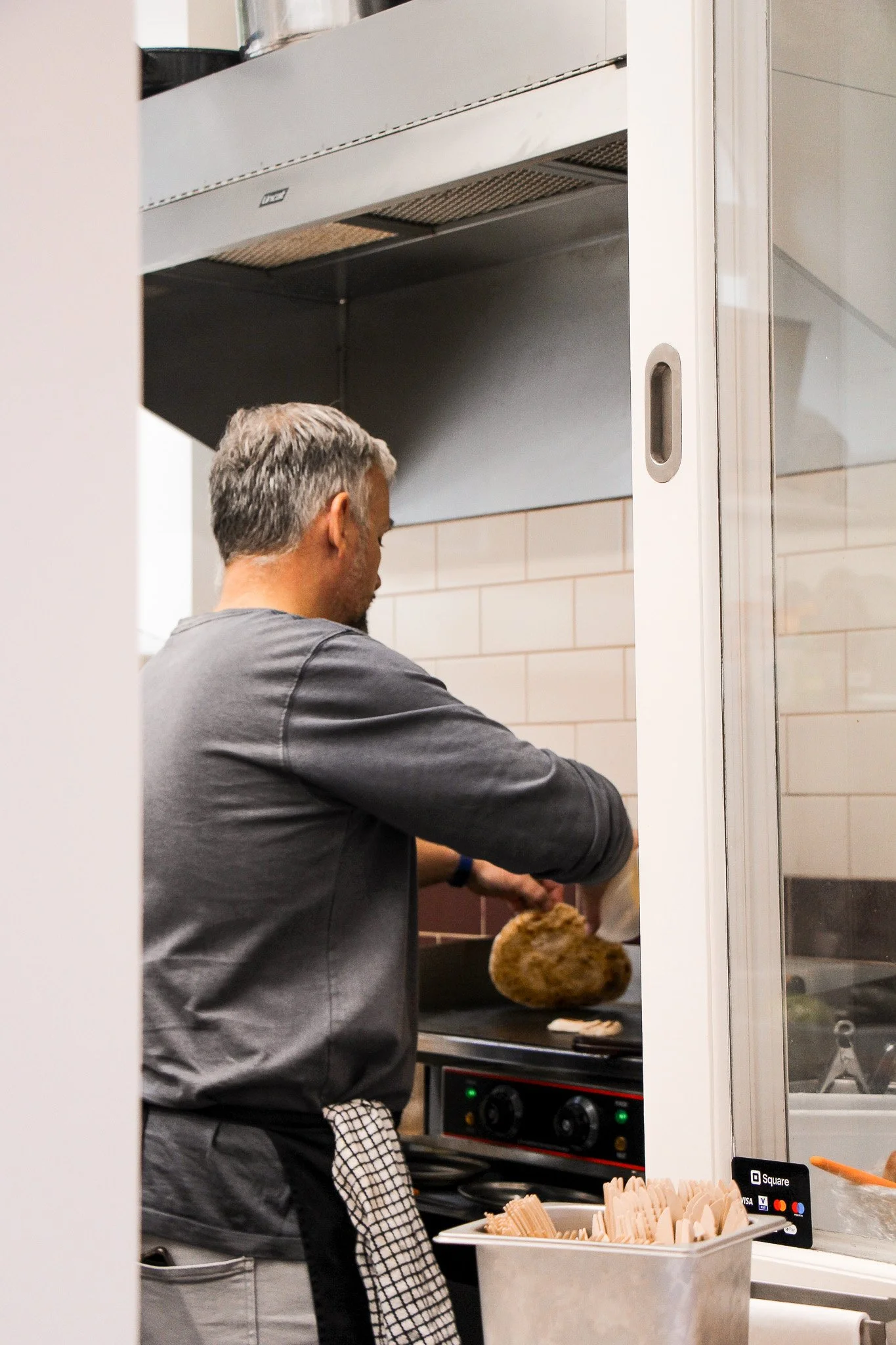 A man working in a kitchen, cooking food over a stove with a metal hood, with a basket of chopsticks and a fried chicken drumstick in the foreground.