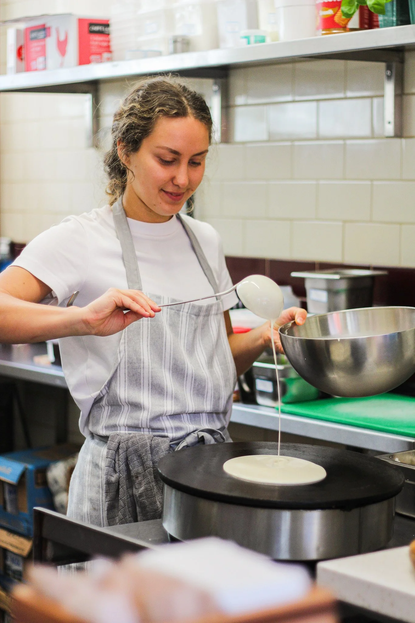 A young woman with curly hair wearing a white shirt and striped apron is cooking on a stovetop, pouring batter onto a hot pan.