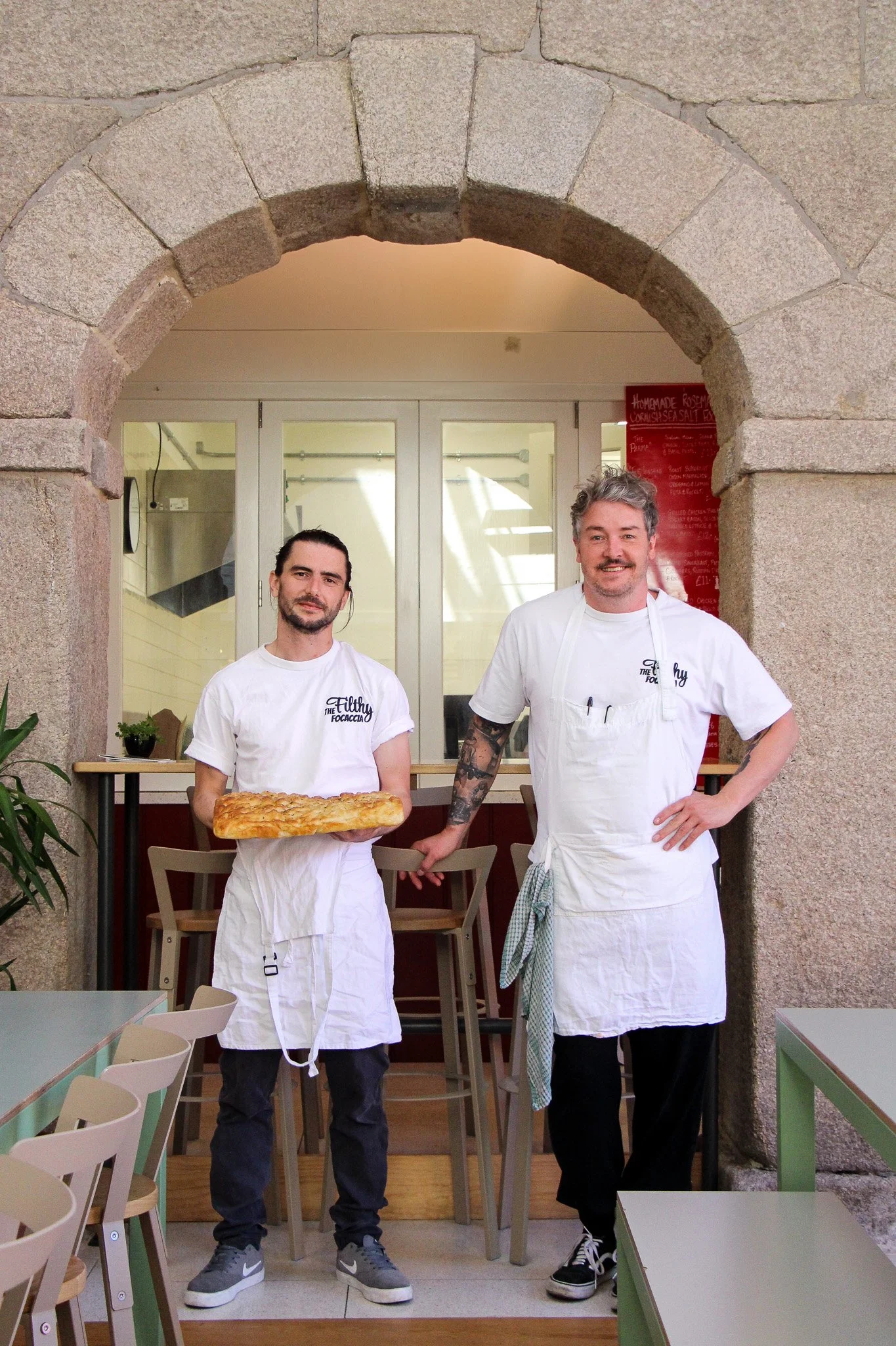 Two male chefs standing in a restaurant kitchen, one holding a pizza and the other wearing a white apron, beneath an arched stone doorway.