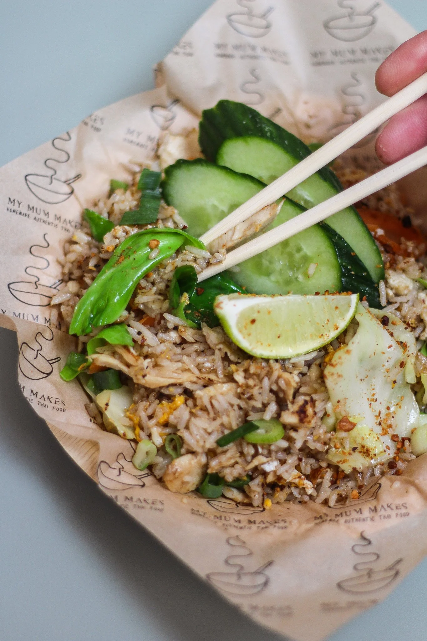 Close-up of fried rice with cucumber slices, green chili, lime wedge, and shredded chicken inside a branded paper bowl with chopsticks.