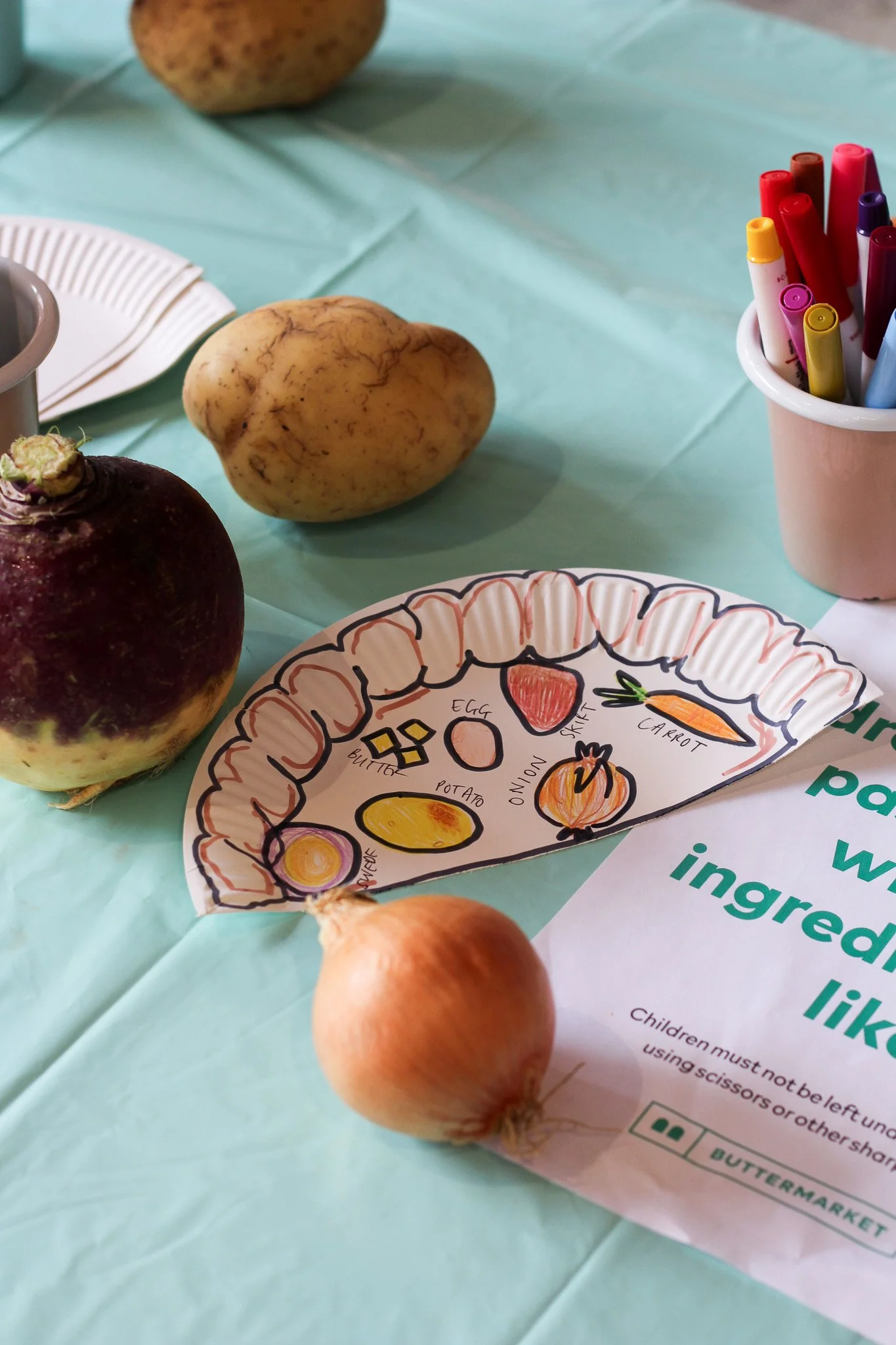A table with potatoes, onions, a paper plate with a drawing of a vegetable tray, a paper cup filled with colored markers, and paper napkins on a light blue tablecloth.