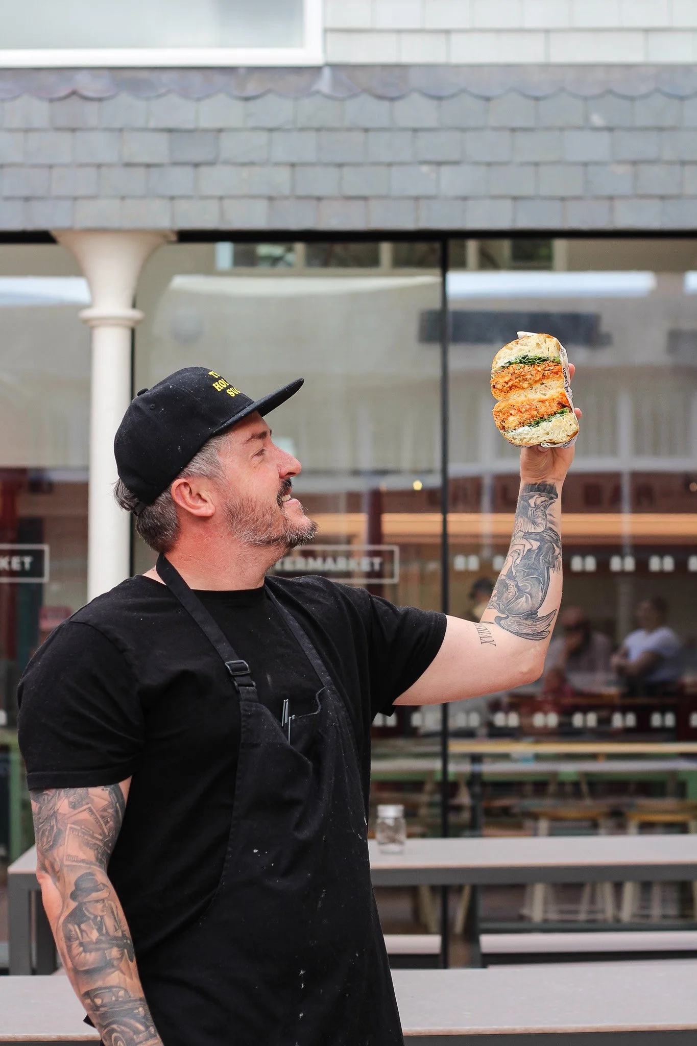 A man with tattoos on his arm, wearing a black cap and black shirt, holding a sandwich in a plastic wrapper, standing outside in front of a building with gray shingles and windows.