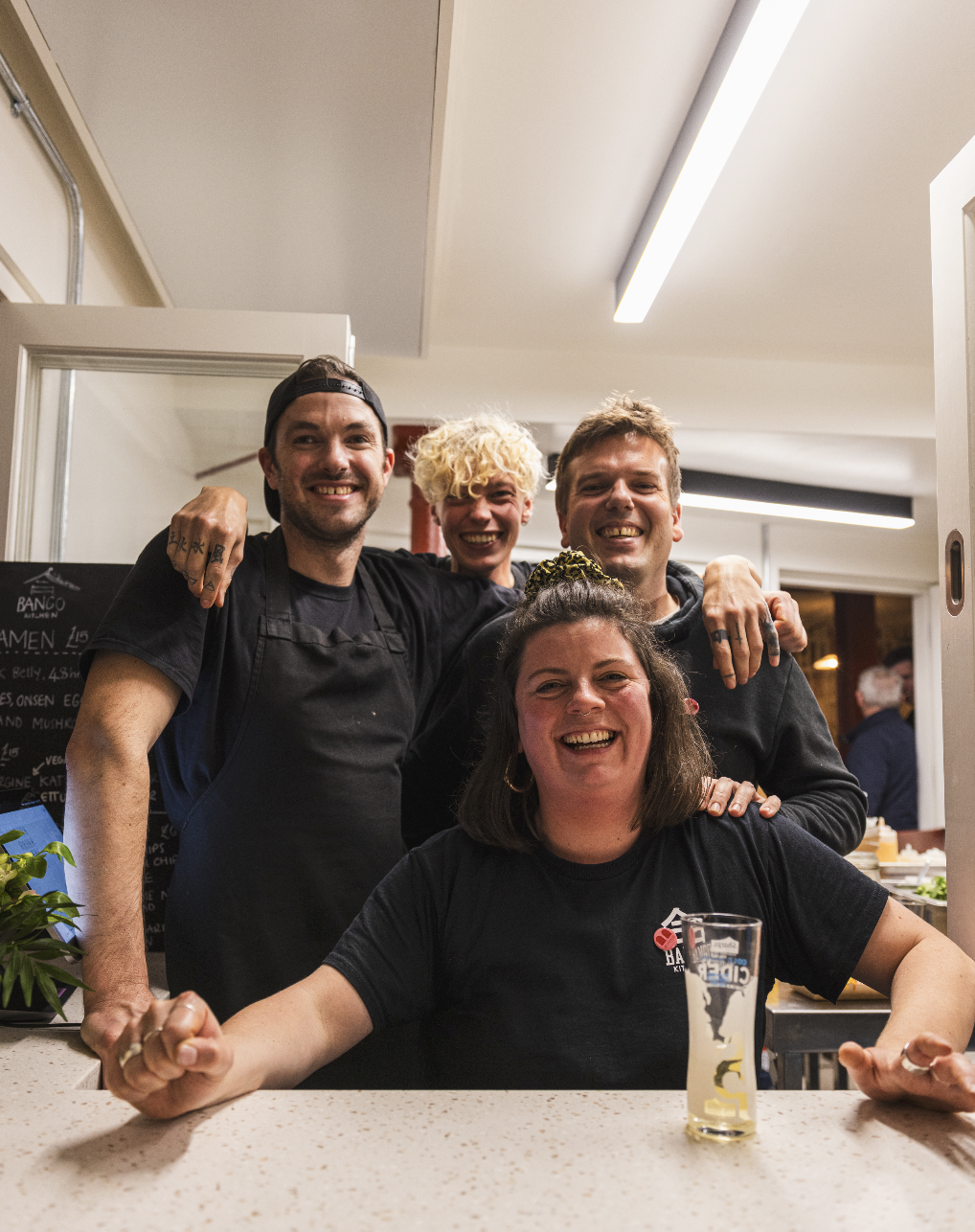 Four people smiling and celebrating at a restaurant bar, with the woman in front having a drink and the group embracing each other.
