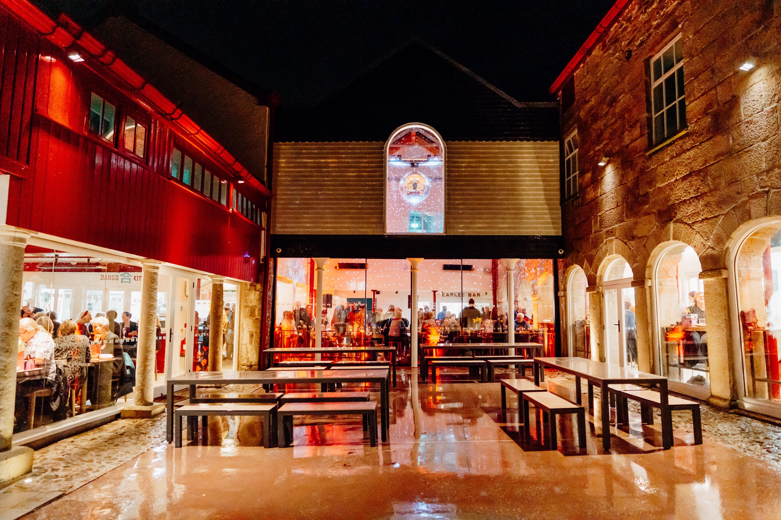 The photo shows the interior of a lively restaurant or food court with many people dining. The decor includes red and beige walls with large windows and an arched window central to the scene. The ceiling is dark, and the floor is reflective with a polished finish. There are tables and benches, and festive or decorative lights are visible through the windows.