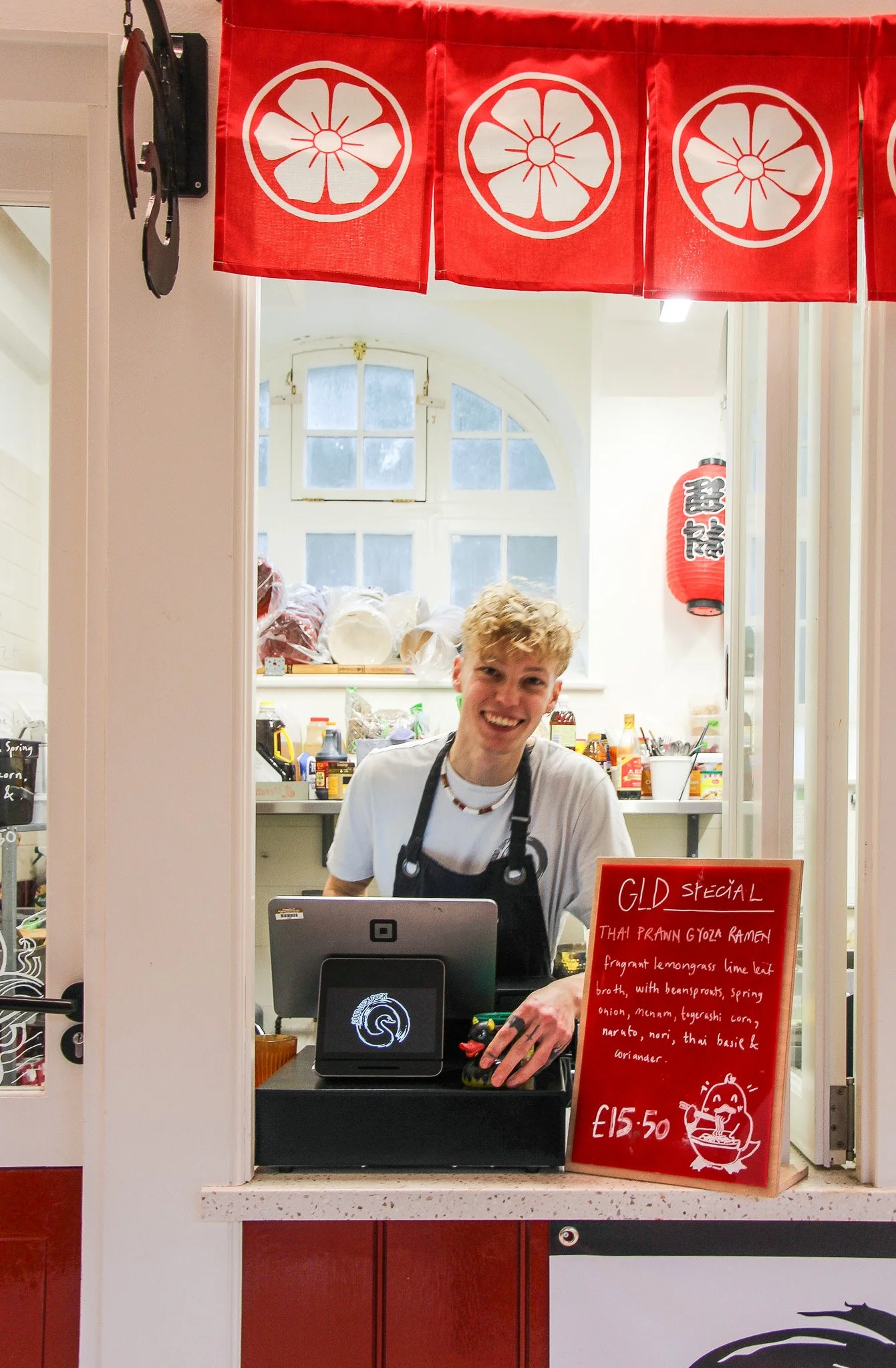 Smiling young man working at a food stand with a red signboard displaying the menu for Thai prawn gyoza ramen, priced at £15.50, with a background of a bright kitchen and decorative red lanterns.