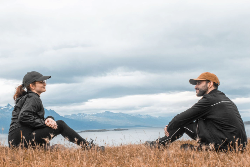 A man and woman sitting on a grassy field near a body of water with mountains in the background. They are facing each other and smiling, dressed in outdoor clothing and caps.
