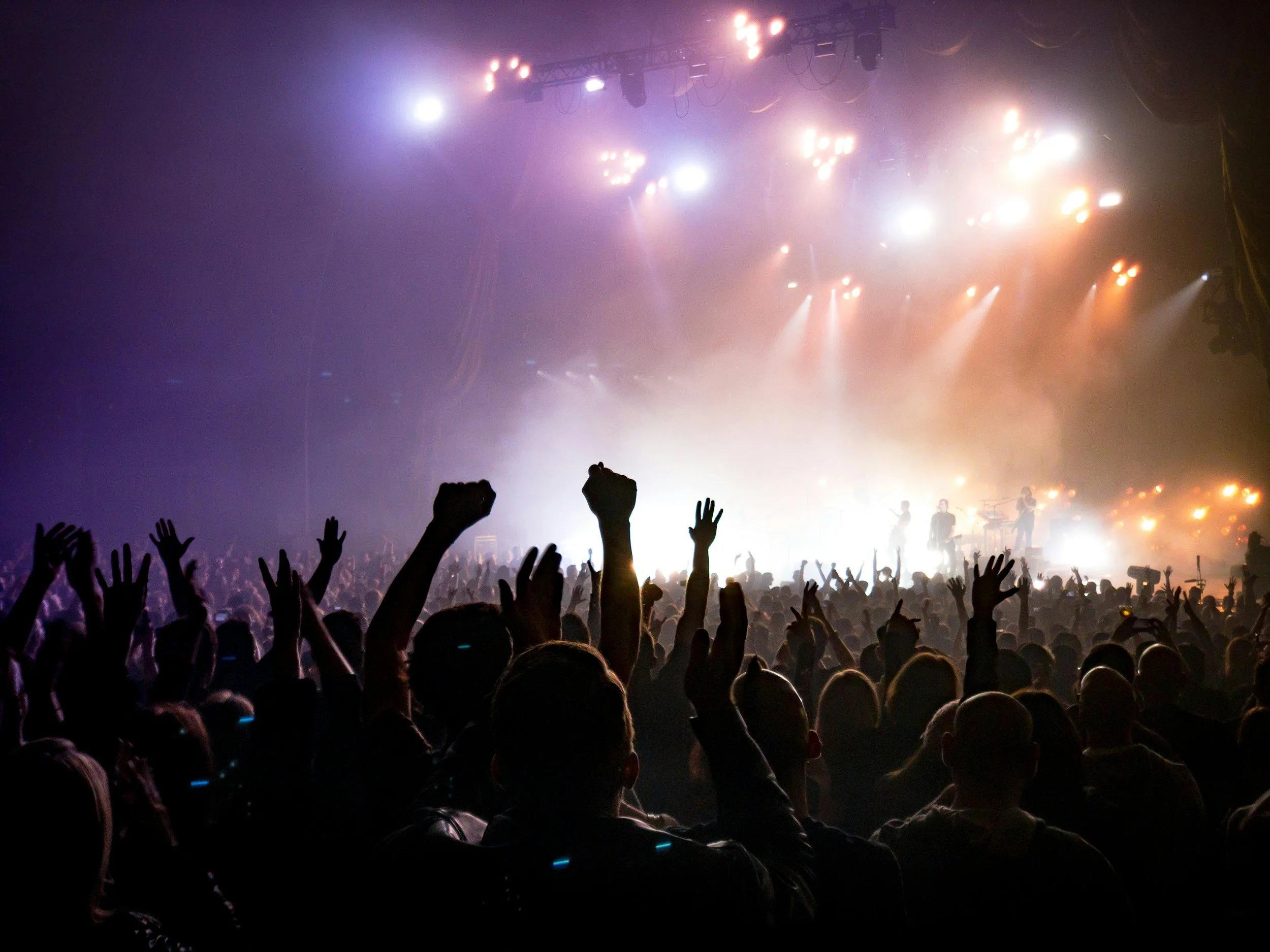 Crowd at a concert with raised hands and a stage with performers and colorful lighting effects.