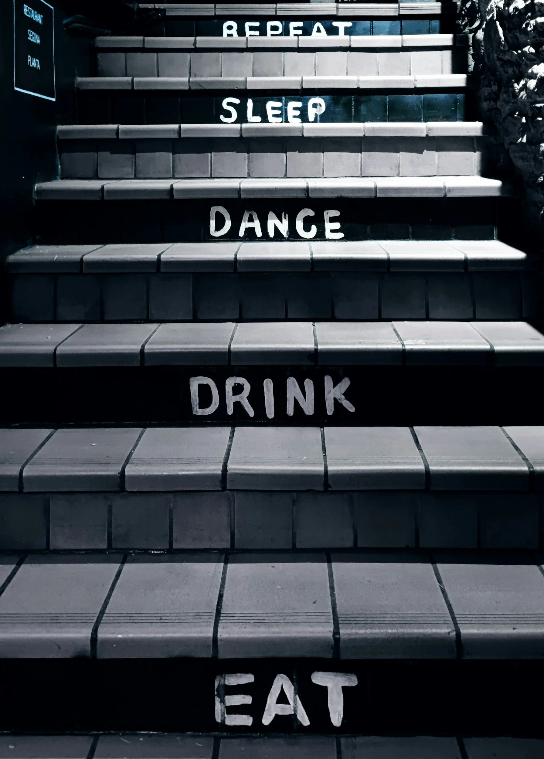 Black-tiled outdoor staircase with white painted words on each step: "REPEAT" on the top step, "SLEEP" on the second step, "DANCE" on the third step, and "EAT" on the bottom step.