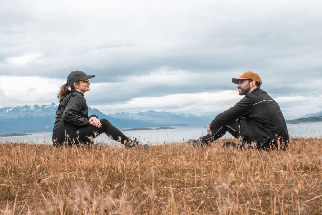 A man and woman sitting on a grassy field near a body of water with mountains in the background. They are facing each other and smiling, dressed in outdoor clothing and caps.