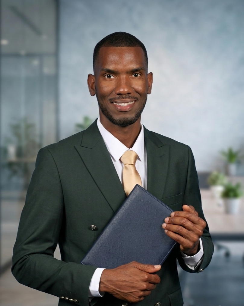 A man in a dark green suit with a cream tie, holding a blue folder, smiling in a modern office setting with plants in the background.