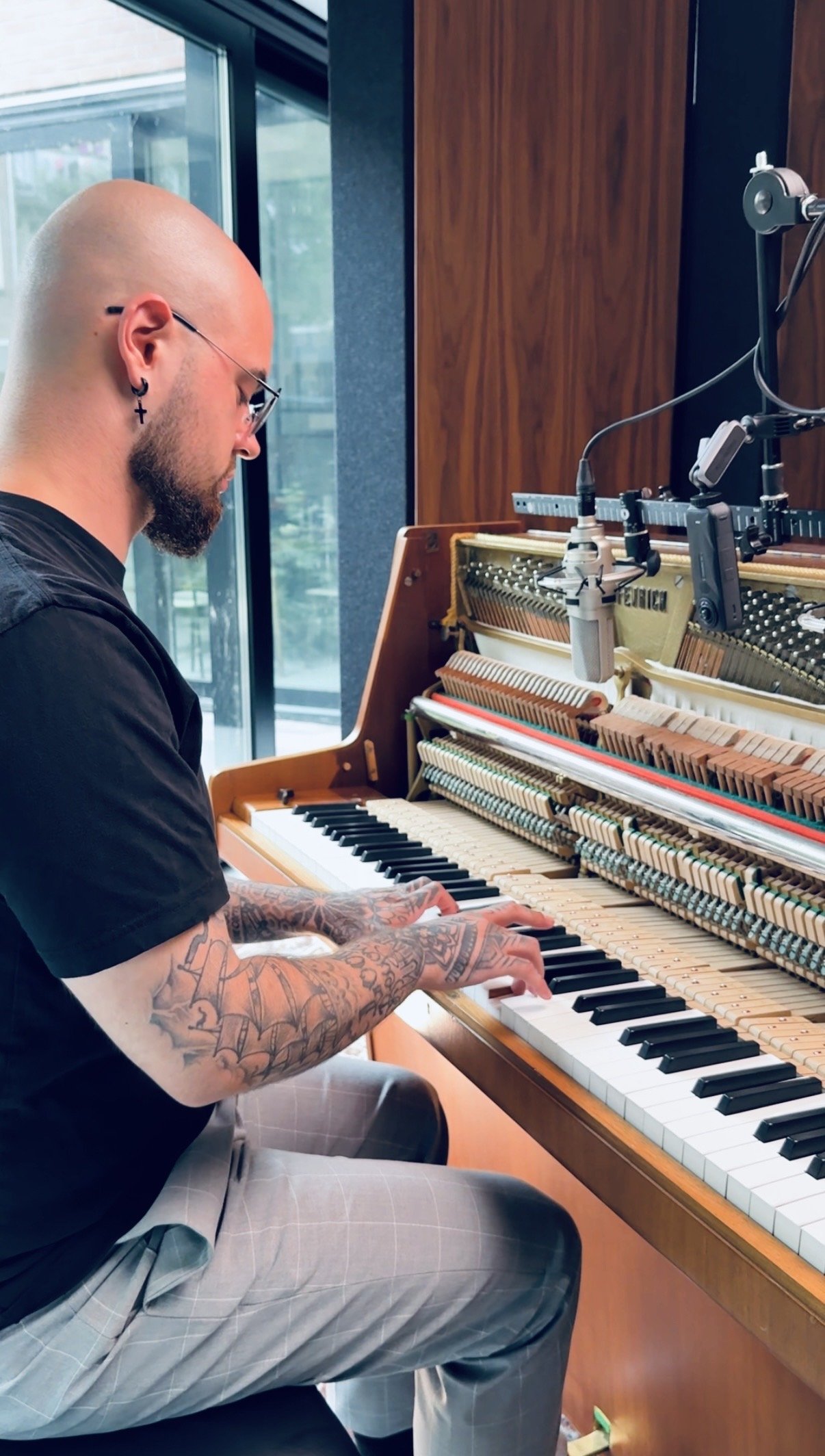 A bald man with tattoos on his arms, wearing glasses and earrings, playing an upright piano with its top open, revealing the internal mechanics, in a room with large windows and wooden walls.