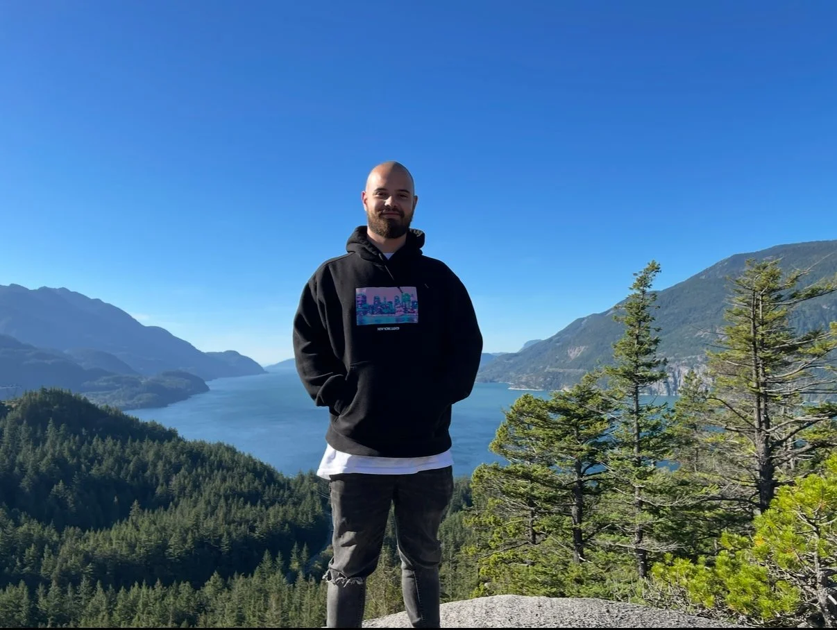 A man standing outdoors on a rocky surface, wearing a black hoodie with a colorful cityscape print, with a body of water and mountains in the background, under a clear blue sky.