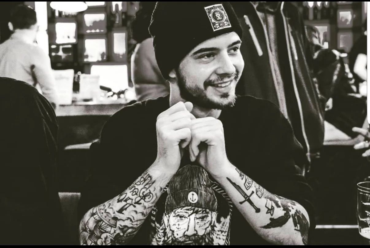 A smiling young man with a beard and tattoos on his arms sits in a restaurant, wearing a black beanie and a dark shirt. The tattoos feature various images including a pirate, a cross, and other designs. There are people and a bar in the blurred background.